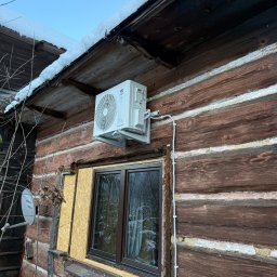 Aleksy Witkowski Kondmaster - Biały klimatyzator na drewnianej ścianie domu z widocznymi przewodami. Zimowy krajobraz z śniegiem na dachu. Widoczna antena satelitarna i zabezpieczone okno.
