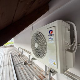 Aleksy Witkowski Kondmaster - Zamontowana jednostka zewnętrzna klimatyzacji Gree Inverter na metalowej konstrukcji wsporczej, widok z dołu, z fragmentem drewnianej konstrukcji dachu.