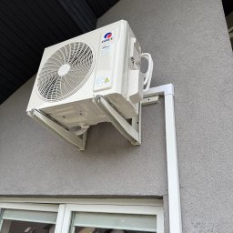 Aleksy Witkowski Kondmaster - Biały klimatyzator marki Gree zamontowany na szarej elewacji budynku, widoczny z dołu, z oknem w tle. Instalacja z rurami odprowadzającymi.