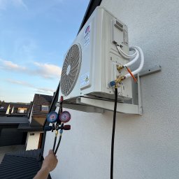 Aleksy Witkowski Kondmaster - Biały klimatyzator GREE zamontowany na elewacji domu, podłączony do manometrów serwisowych. Widoczne przewody i uchwyt montażowy. Ujęcie z dołu, niebo w tle.