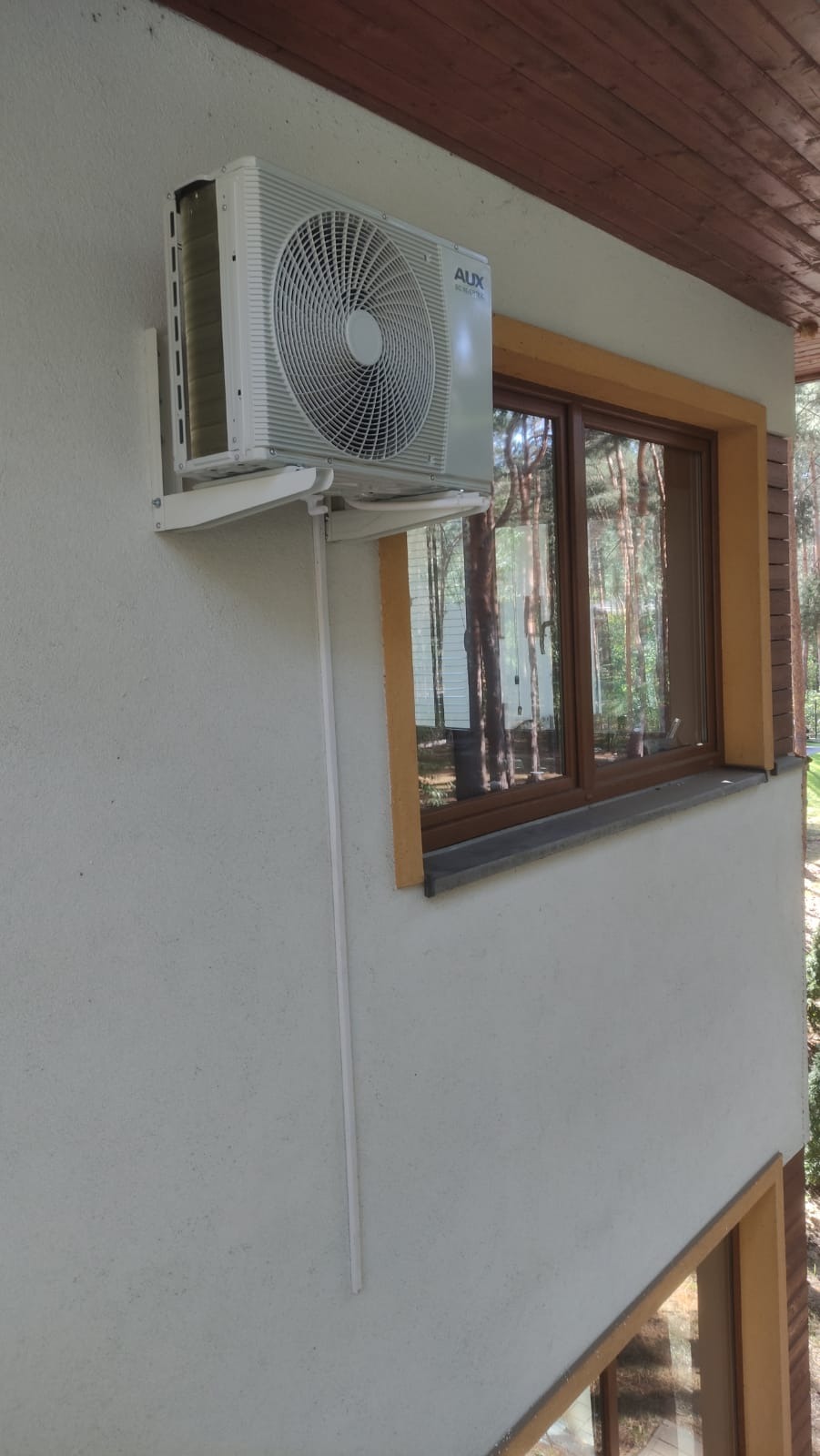 Zewnętrzna jednostka klimatyzacji AUX zamontowana na elewacji domu obok okna z brązowymi ramami. Widoczna biała listwa maskująca instalację.