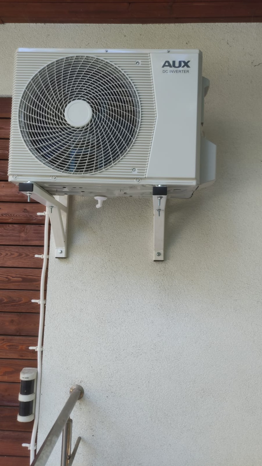 Zewnętrzna jednostka klimatyzatora AUX DC Inverter zamontowana na białej ścianie z drewnianą okładziną w tle, widoczne wsporniki mocujące i przewody.
