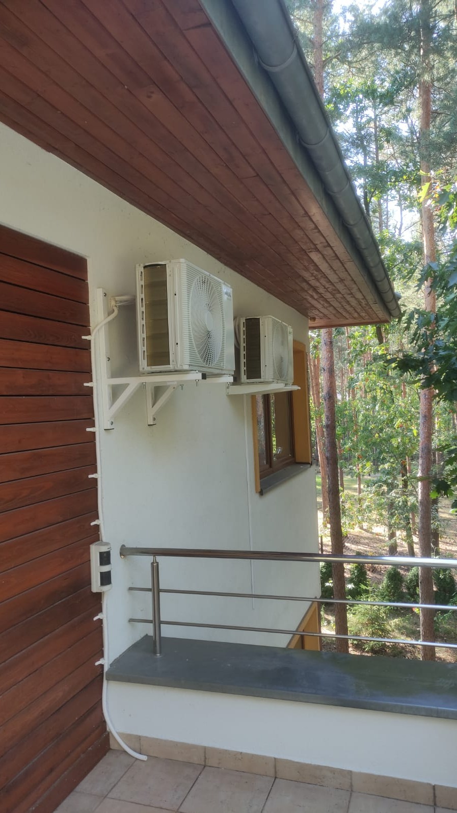 Dwa białe klimatyzatory zamontowane na wspornikach na ścianie budynku z drewnianą elewacją i brązowym podbitką dachową, widoczne drzewa w tle.
