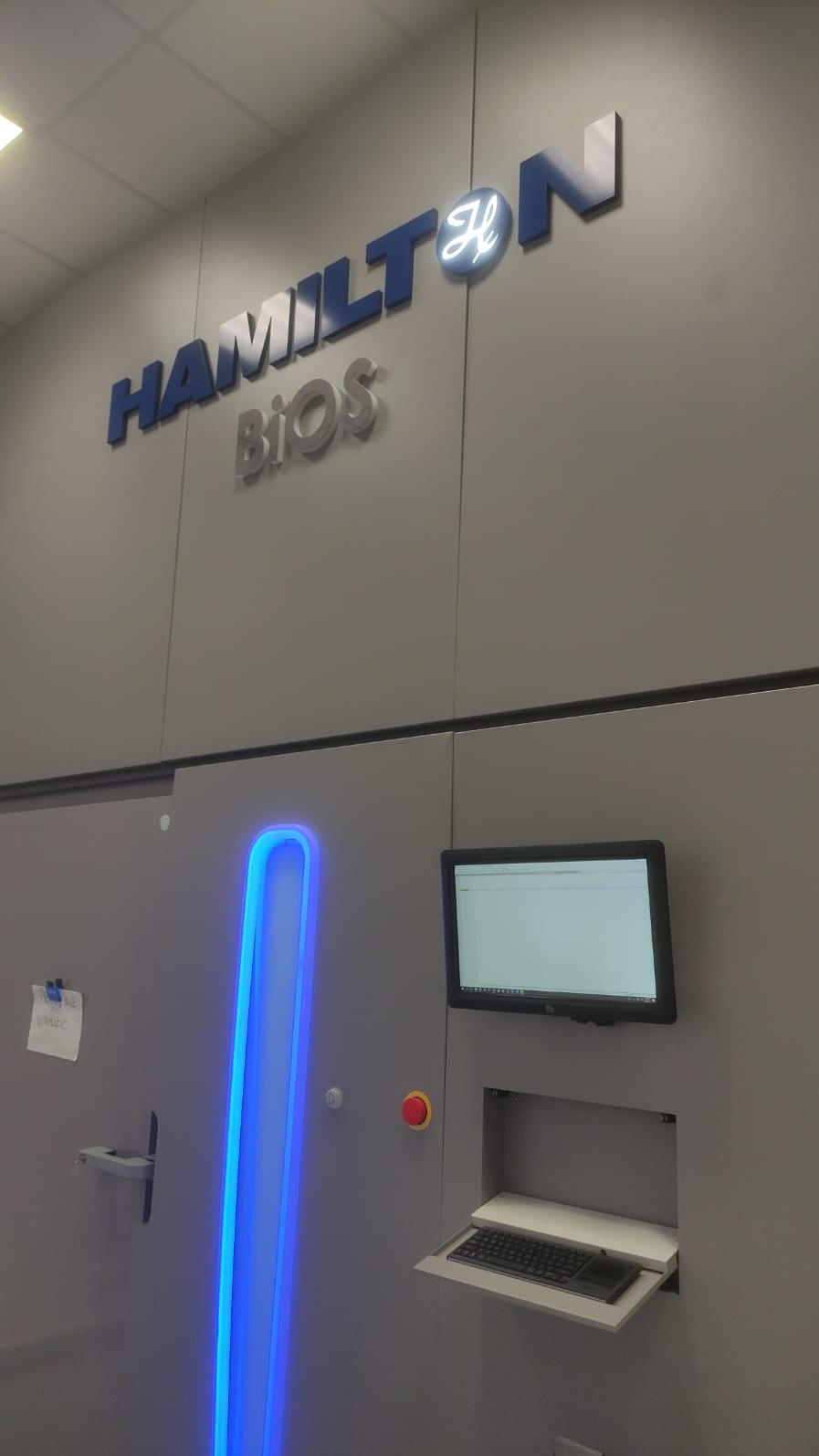 Szara ściana z podświetlonym logo Hamilton Bios, obok panel sterowania z monitorem i wysuwaną klawiaturą.