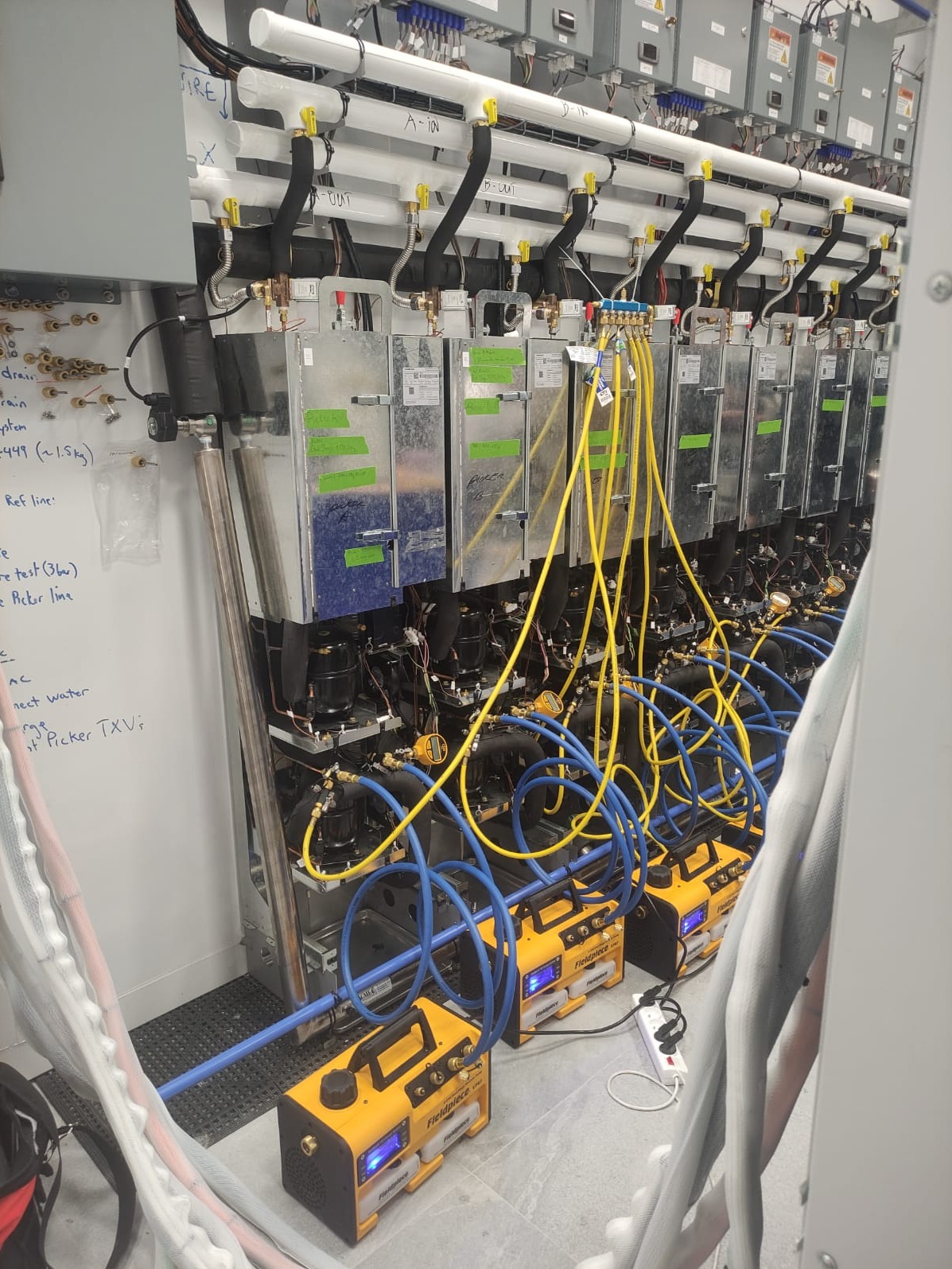 Serwis klimatyzacji w Spytkowicach: Rząd jednostek klimatyzacyjnych podłączonych do pomp próżniowych Fieldpiece za pomocą niebieskich i żółtych przewodów, widoczne notatki na ścianie.