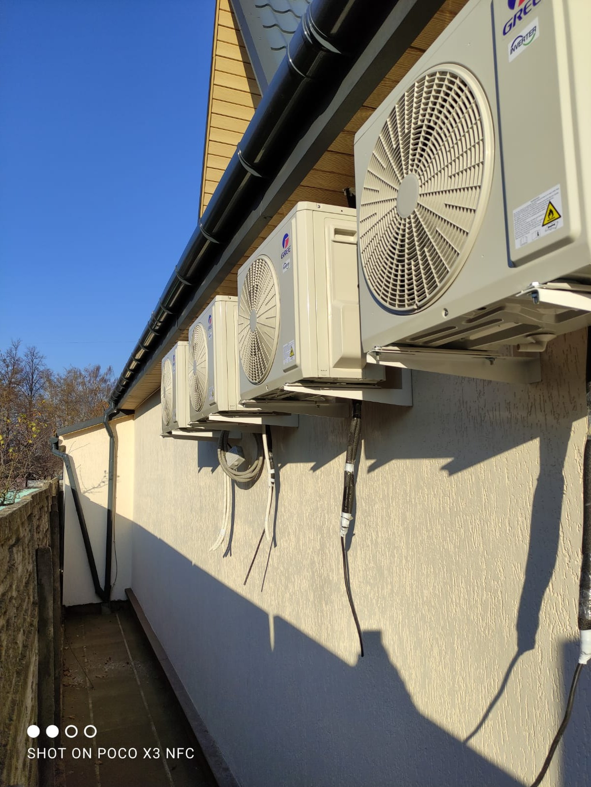 Rząd jednostek zewnętrznych klimatyzacji marki Gree zamontowanych na ścianie budynku z widocznymi przewodami i rynną dachową w słoneczny dzień.