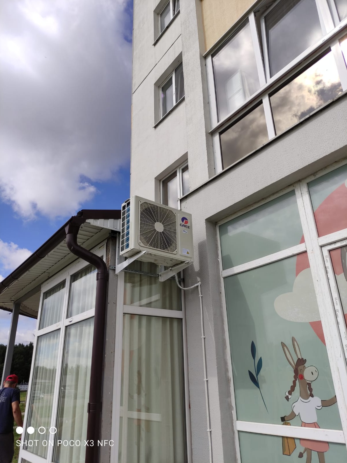 Zewnętrzny moduł klimatyzacji marki Gree zamontowany na elewacji budynku z oknami, widoczne rury odprowadzające skropliny.