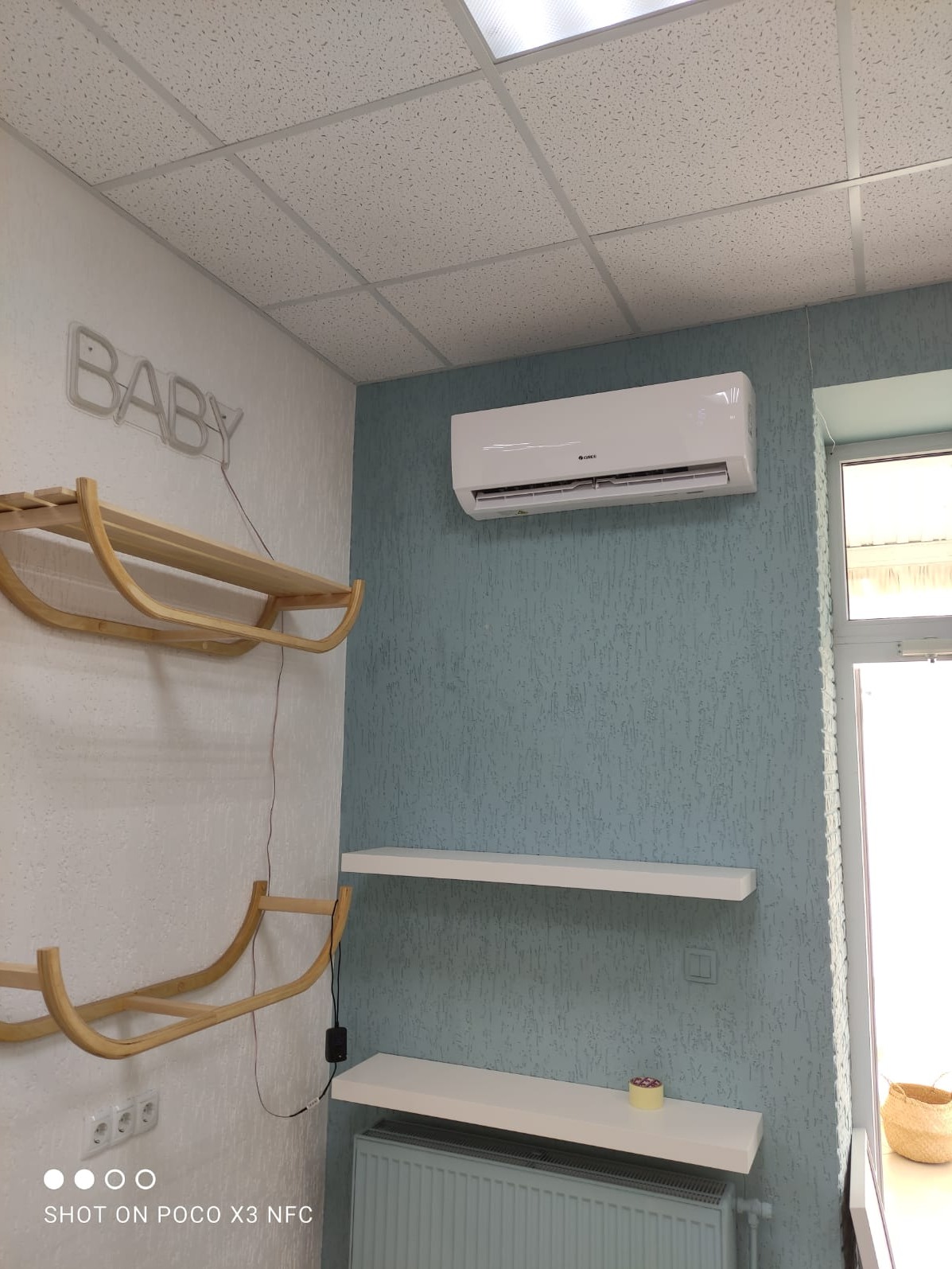 Biały klimatyzator zamontowany na błękitnej ścianie w pokoju dziecięcym, pod nim białe półki i grzejnik, po lewej stronie dekoracja ścienna w kształcie sanek i neon 'BABY'.