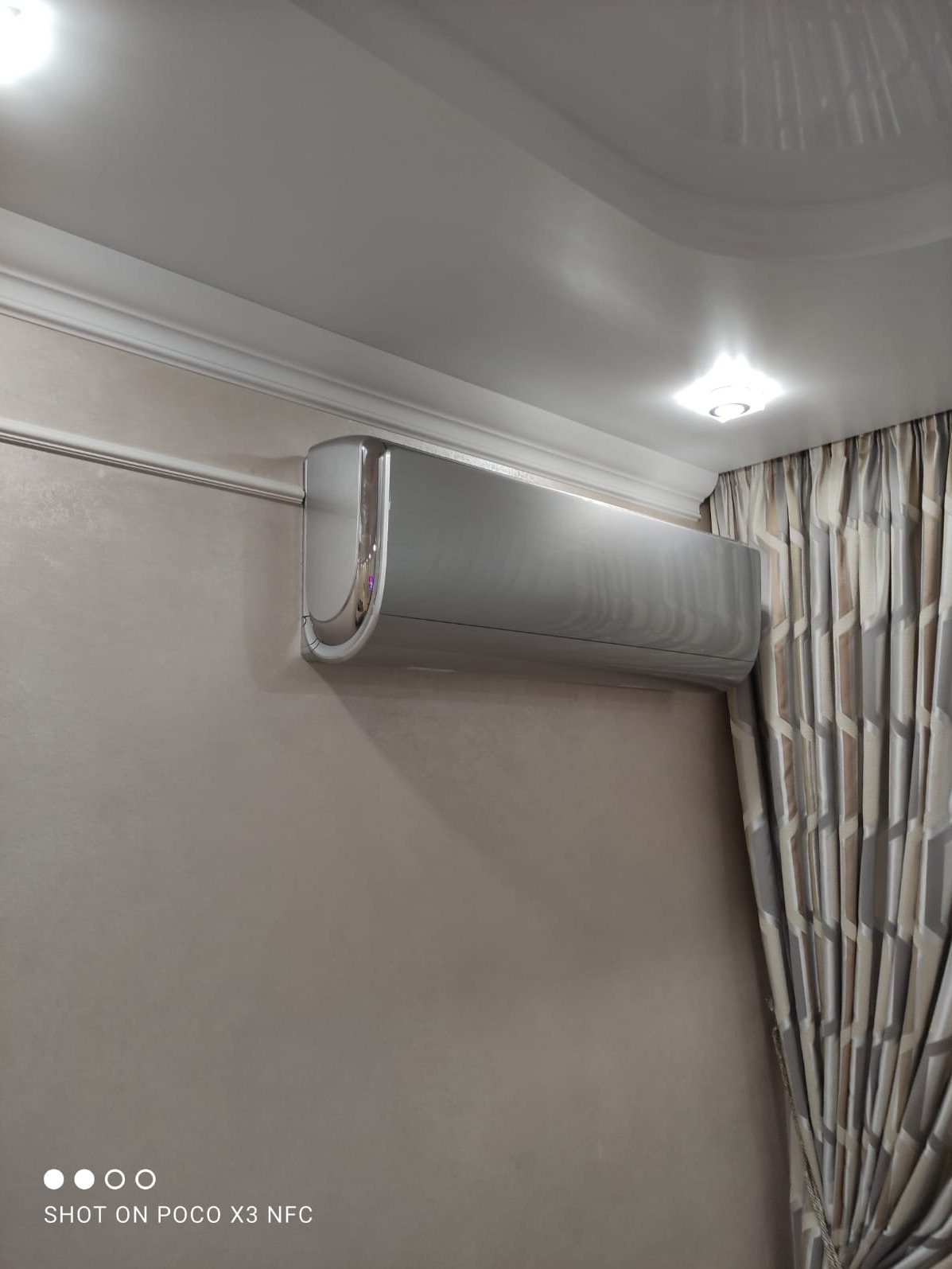 Nowoczesny, srebrny klimatyzator ścienny w jasnym wnętrzu, z widocznym fragmentem sufitu z listwą dekoracyjną, oświetleniem i zasłonami w geometryczne wzory.