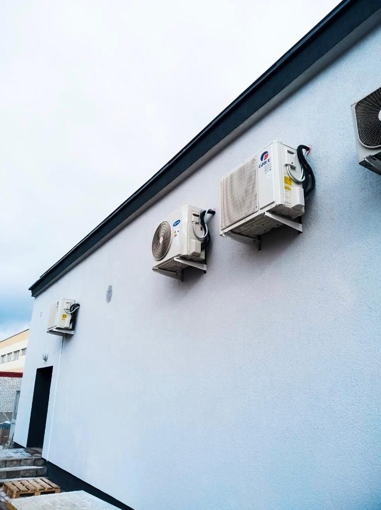 Biała ściana budynku z trzema zamontowanymi jednostkami zewnętrznymi klimatyzacji marki Gree, widoczne przewody i wsporniki montażowe, pochmurne niebo w tle.