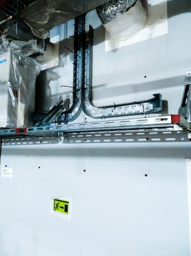 Metalowe korytka kablowe zamontowane pod sufitem, z widoczną instalacją klimatyzacji w folii ochronnej i znakiem ewakuacyjnym na ścianie.