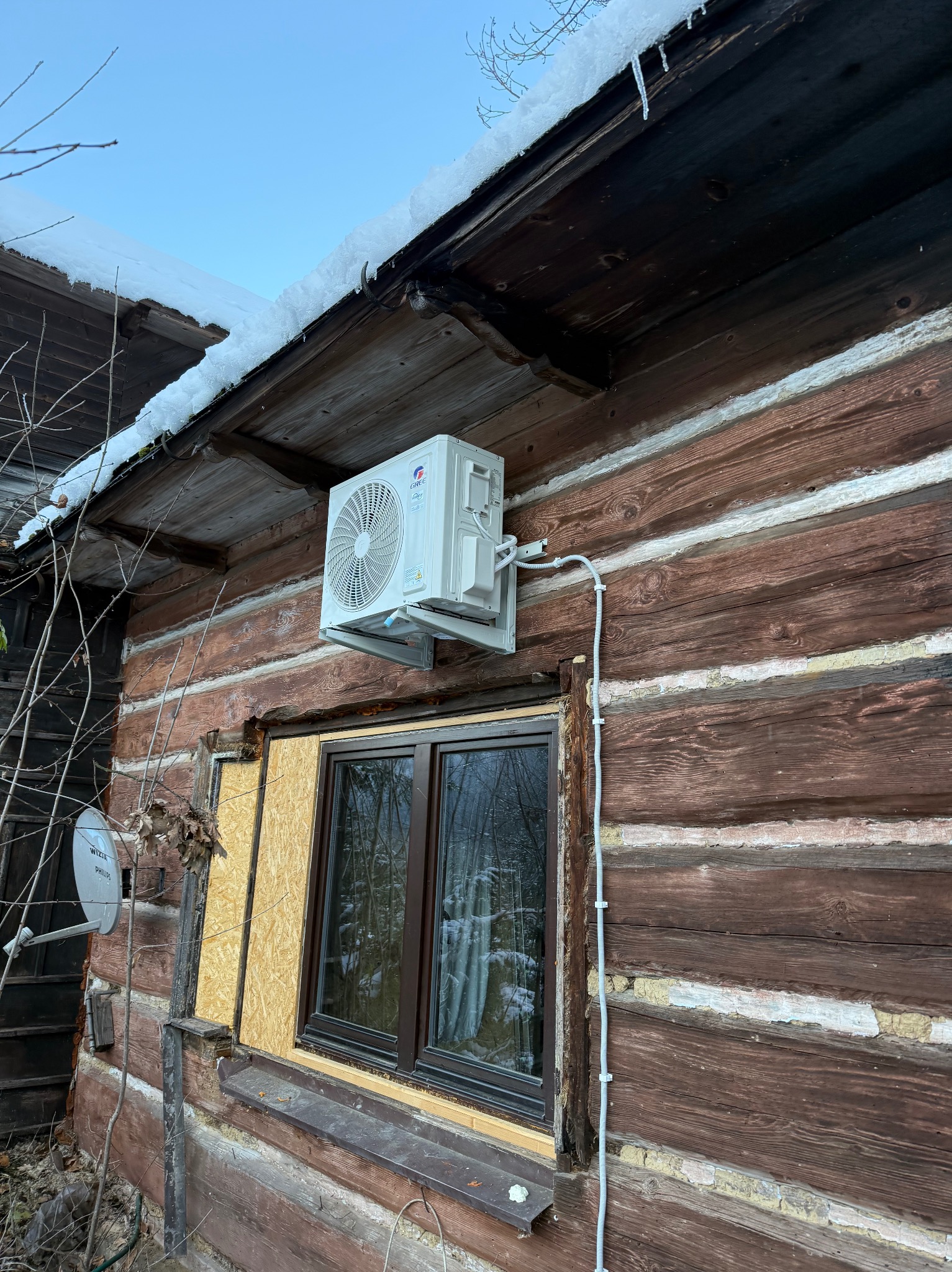 Biały klimatyzator na drewnianej ścianie domu z widocznymi przewodami. Zimowy krajobraz z śniegiem na dachu. Widoczna antena satelitarna i zabezpieczone okno.