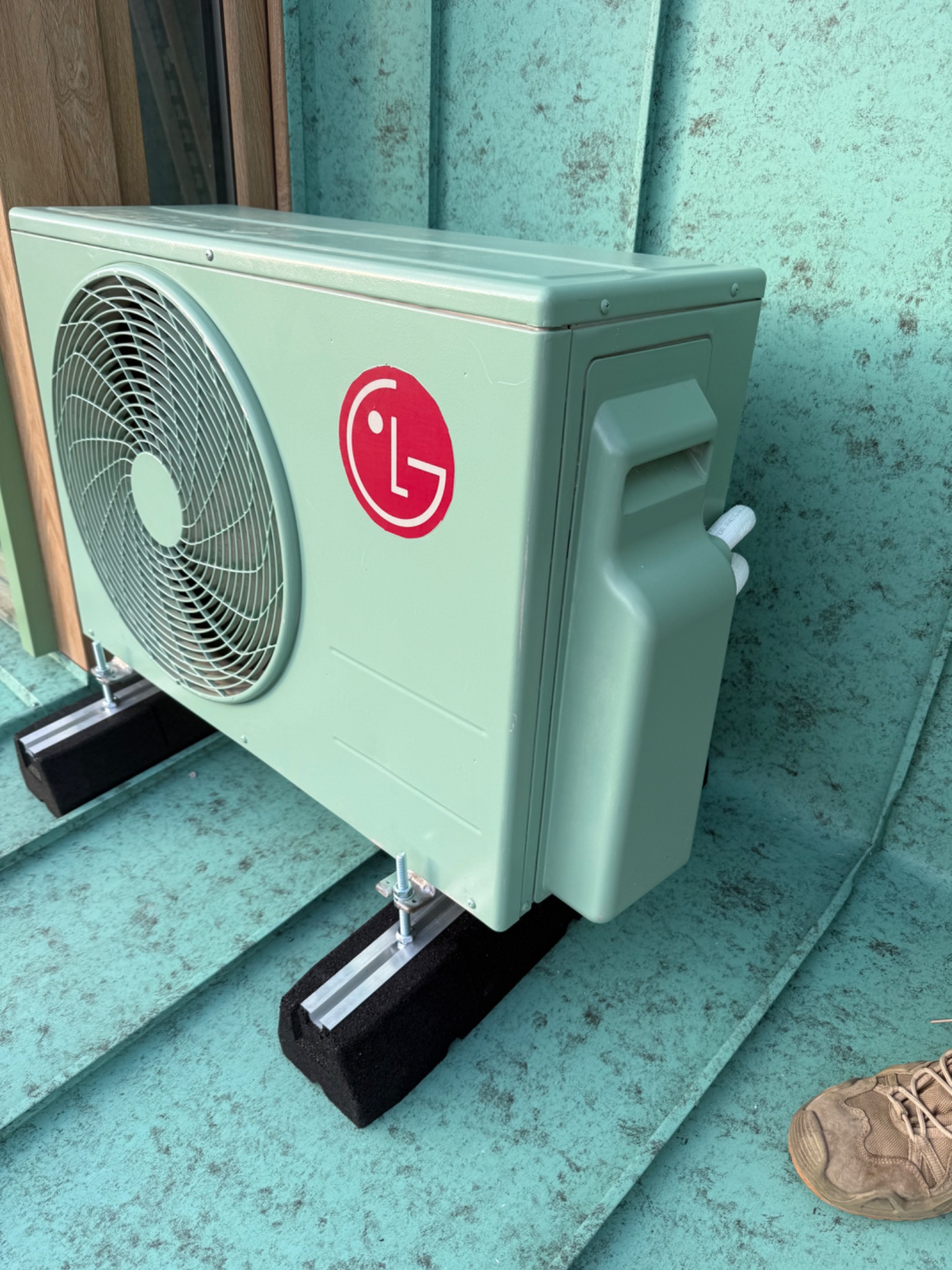 Jasnozielony klimatyzator LG zamontowany na dachu z blachy w kolorze morskim, widoczne czarne podkładki antywibracyjne i fragment buta. Nietypowa kolorystyka.