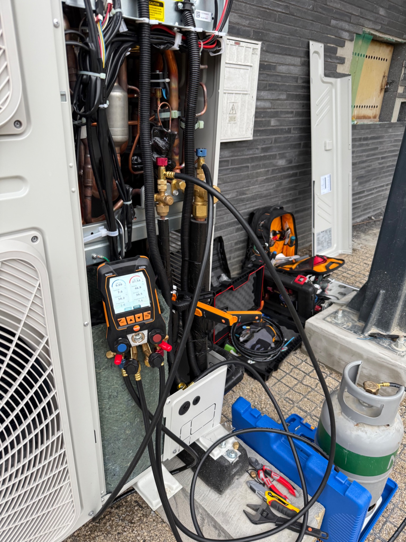 Otwarta obudowa klimatyzatora z widocznymi przewodami, rurami i podłączonym miernikiem ciśnienia. Obok narzędzia i butla z gazem chłodniczym.