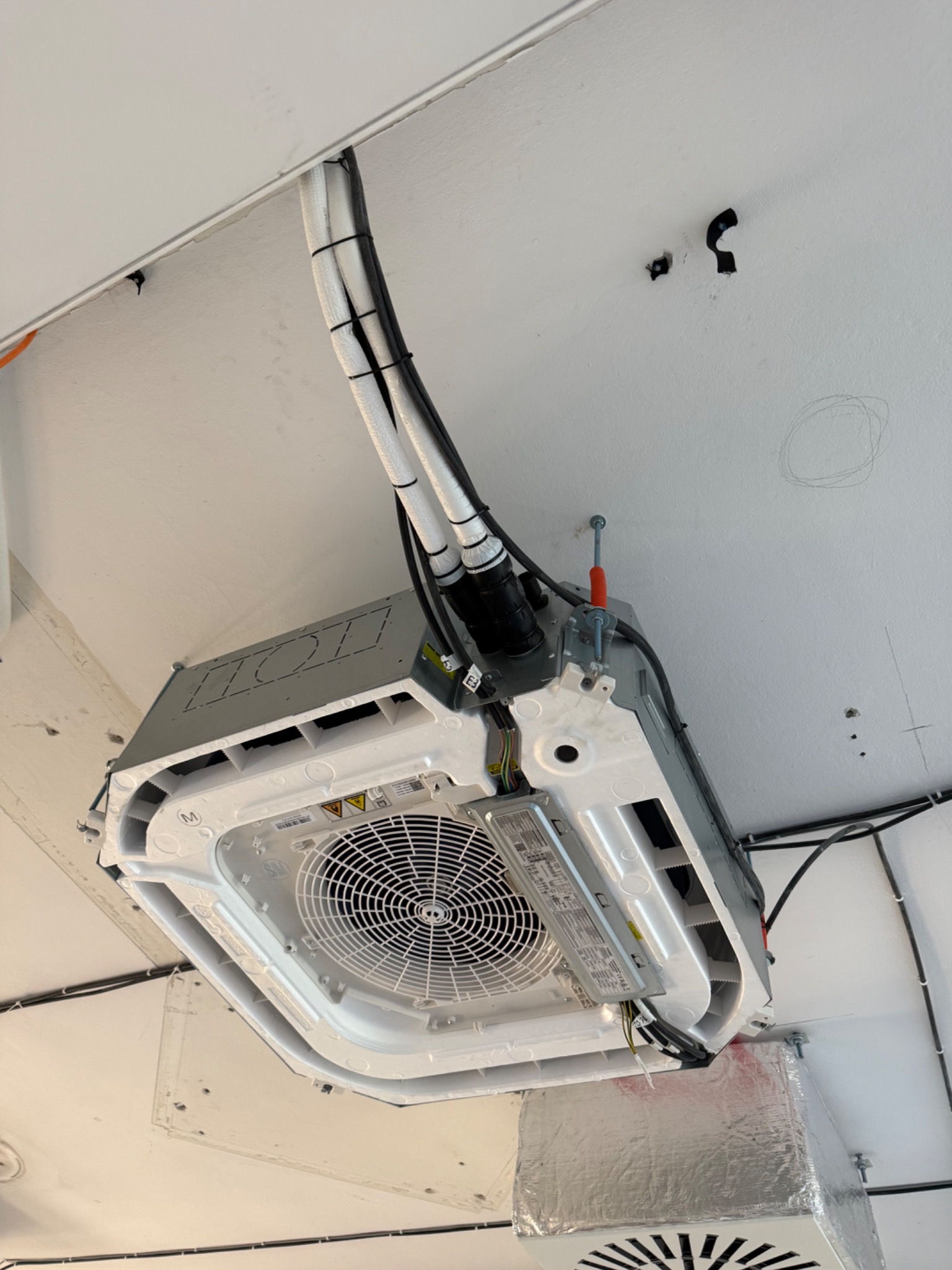 Sufitowa jednostka klimatyzacji kasetonowej z widocznymi przewodami i elementami montażowymi, zamontowana na białym suficie. Widok z dołu, z bliska.