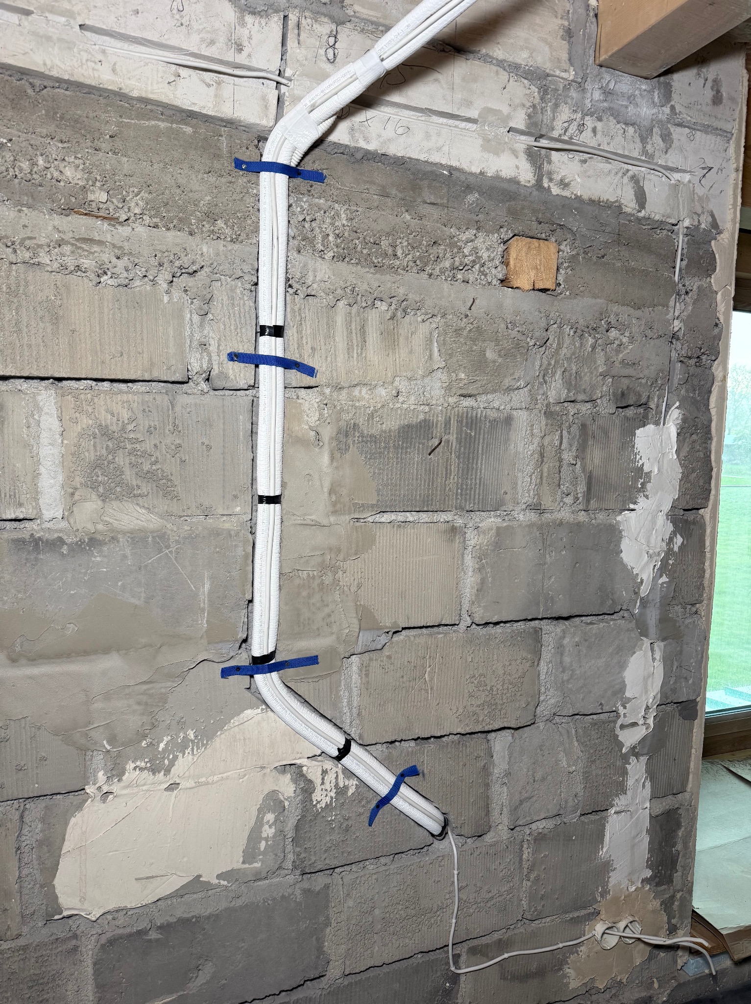 Białe przewody klimatyzacji przymocowane do surowej ściany z betonu komórkowego za pomocą niebieskich opasek. Widoczne ślady po kuciu i naprawy tynkarskie.