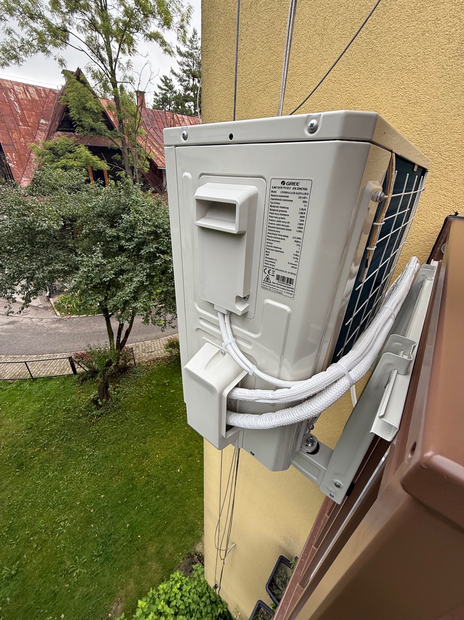 Zewnętrzna jednostka klimatyzacji Gree zamontowana na żółtej ścianie budynku, widok z góry na trawnik i drzewo. Widoczne białe przewody i uchwyty montażowe.