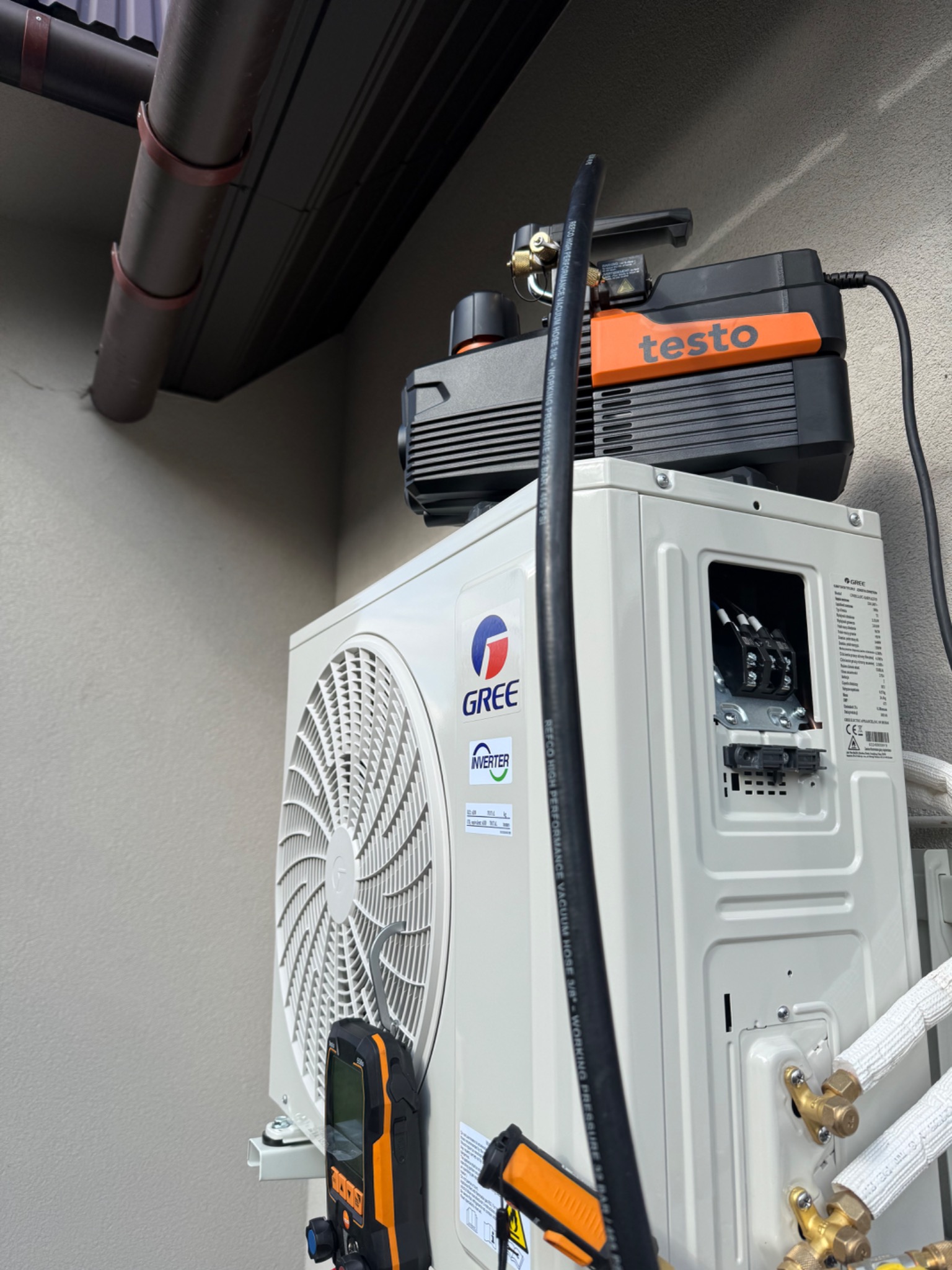 Biała jednostka zewnętrzna klimatyzacji GREE z podłączoną pompą próżniową Testo i manometrem. Widoczne rury chłodnicze i przewody elektryczne. Montaż lub serwis klimatyzacji.