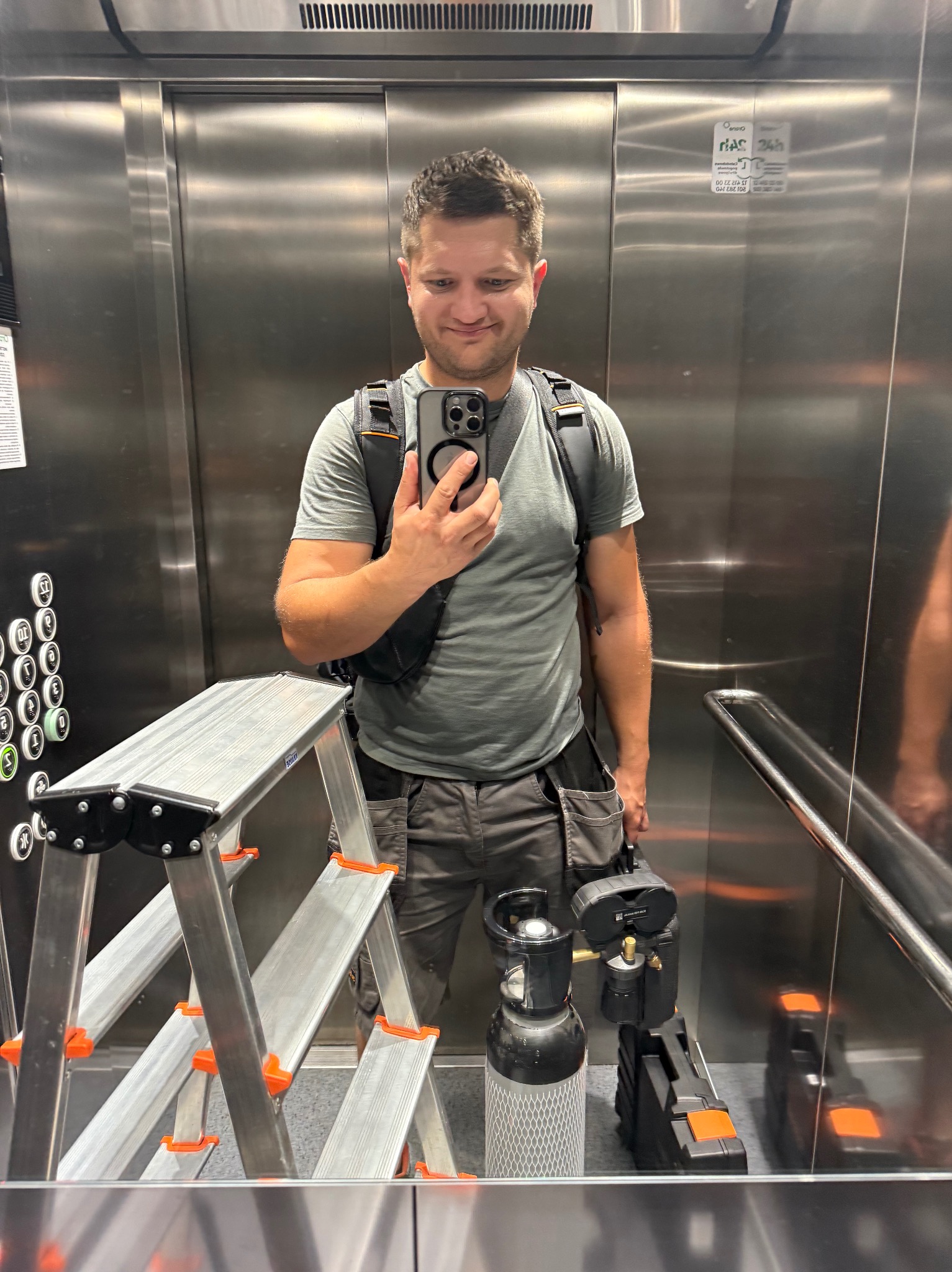 Technik w windzie z butlą gazową i narzędziami do klimatyzacji, robiący selfie. Widoczna drabina i przyciski windy. Metalowe wykończenie wnętrza.