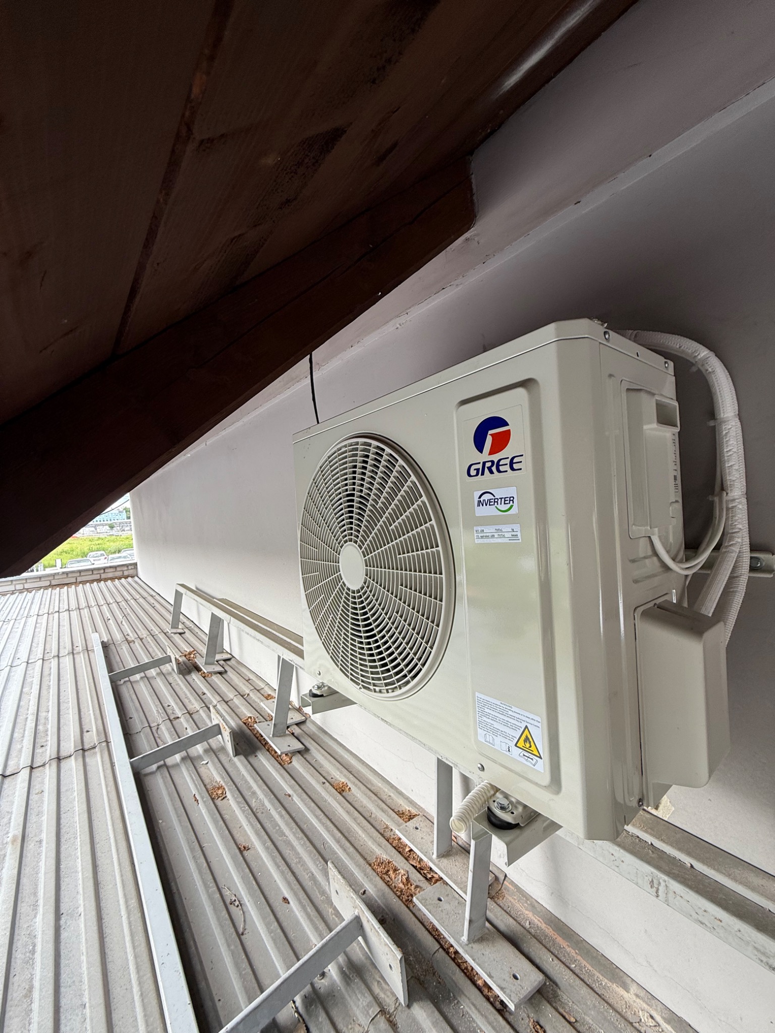 Zamontowana jednostka zewnętrzna klimatyzacji Gree Inverter na metalowej konstrukcji wsporczej, widok z dołu, z fragmentem drewnianej konstrukcji dachu.