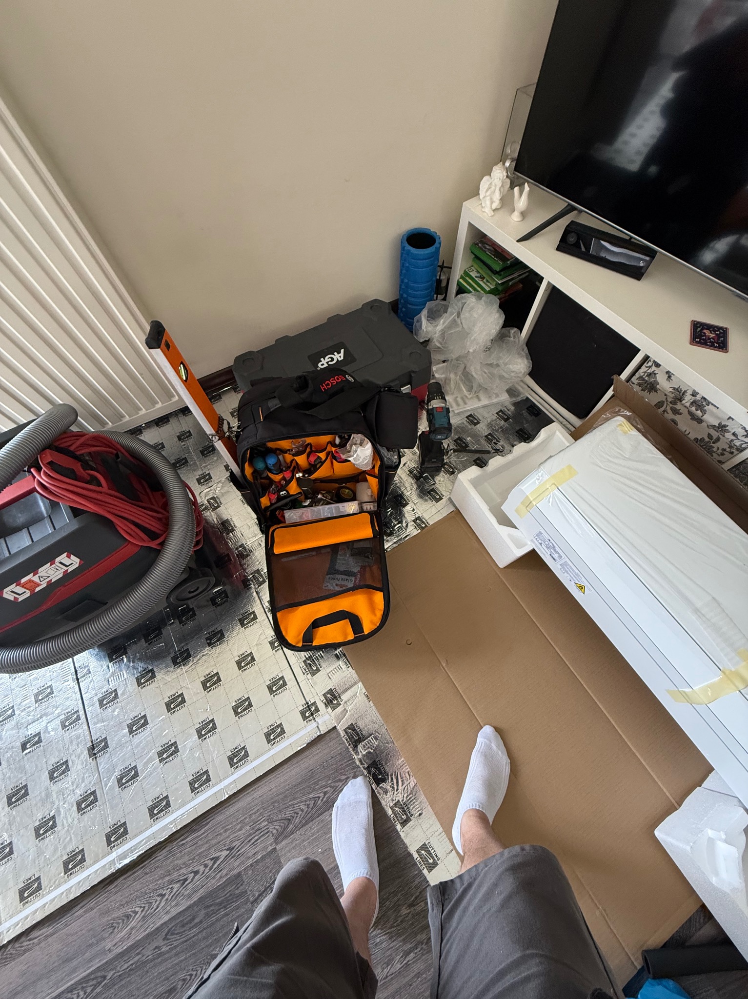 Montaż klimatyzacji w domu: rozpakowane urządzenie, torba z narzędziami Bosch, poziomica i odkurzacz na podłodze wyłożonej folią izolacyjną. Widok z góry, z nogami instalatora.