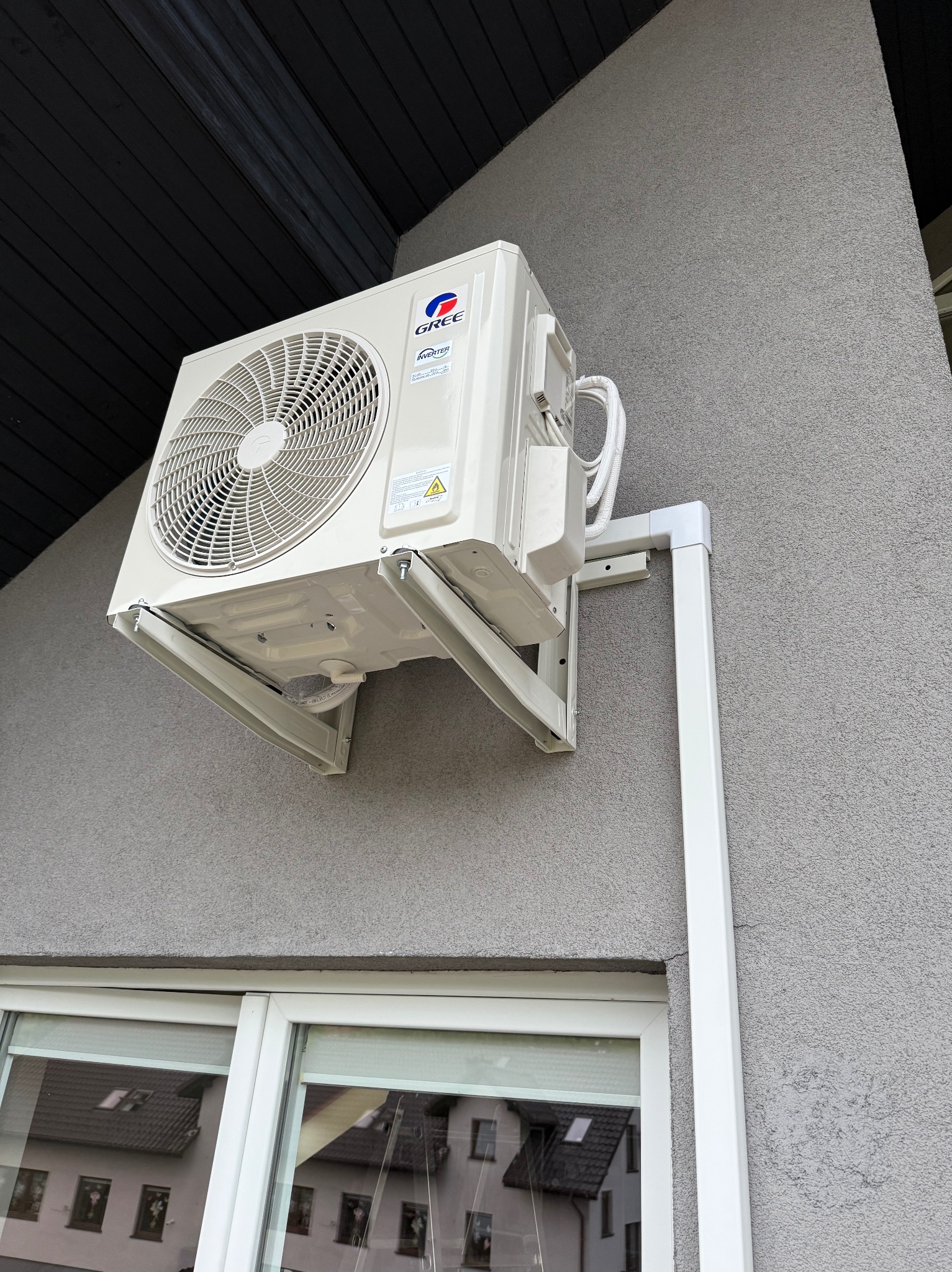 Biały klimatyzator marki Gree zamontowany na szarej elewacji budynku, widoczny z dołu, z oknem w tle. Instalacja z rurami odprowadzającymi.