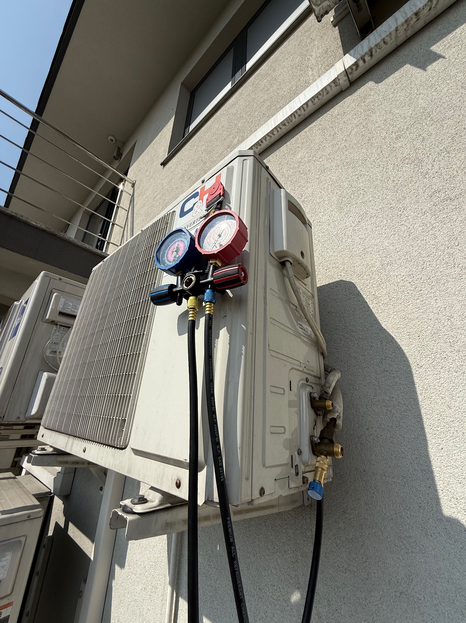 Podłączenie manometrów do klimatyzatora zewnętrznego na elewacji budynku. Widoczne przewody i zawory. Diagnostyka klimatyzacji w trakcie serwisu.