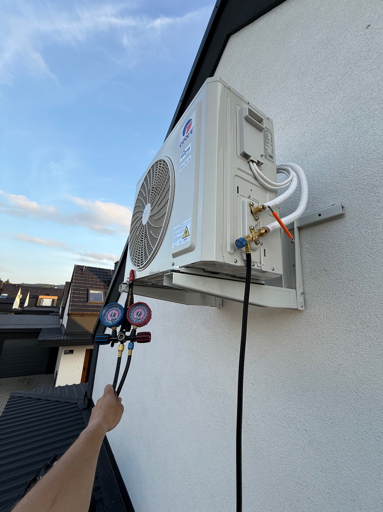 Biały klimatyzator GREE zamontowany na elewacji domu, podłączony do manometrów serwisowych. Widoczne przewody i uchwyt montażowy. Ujęcie z dołu, niebo w tle.