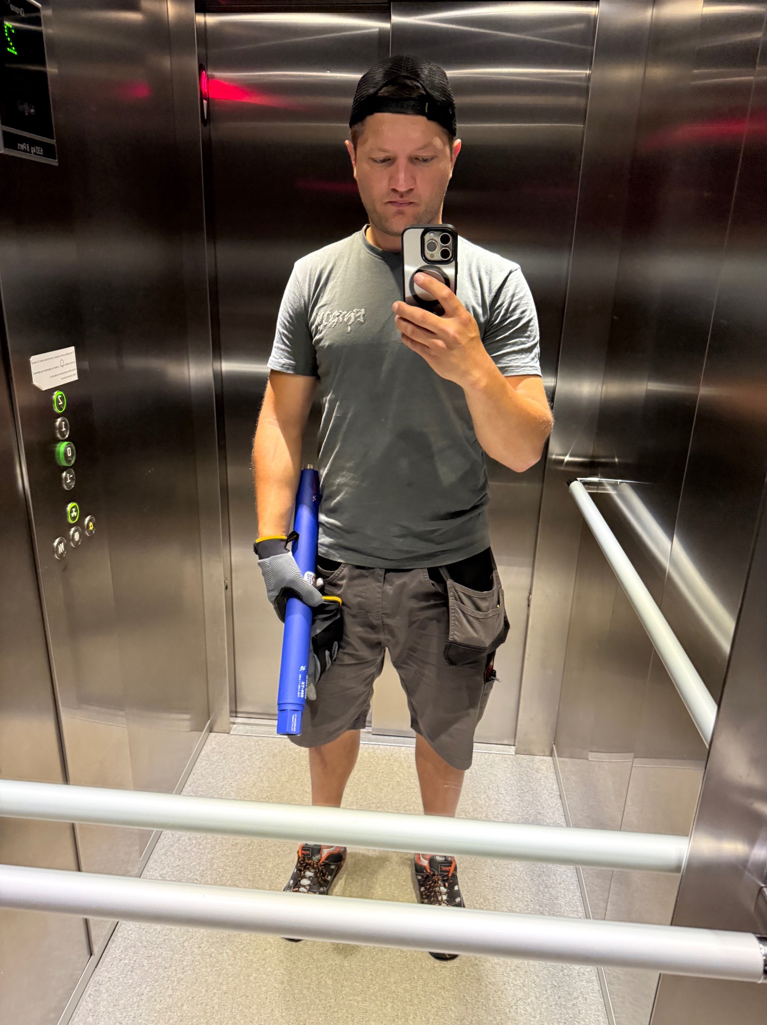 Mężczyzna w windzie, ubrany w robocze ubranie, robi selfie trzymając niebieski element klimatyzacji. Widoczne narzędzia w kieszeniach i przyciski windy.