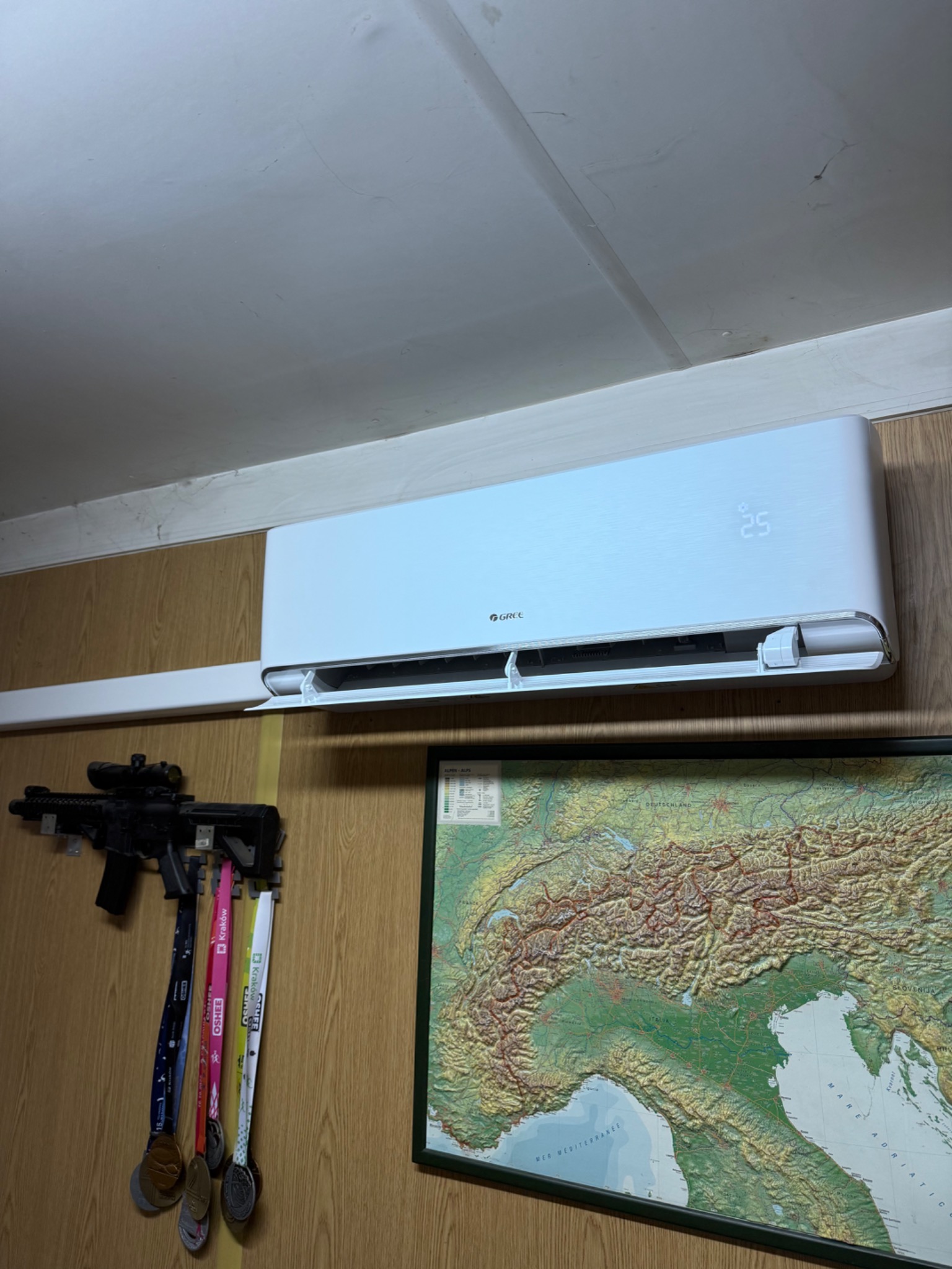 Biały klimatyzator Gree na ścianie z boazerią, obok mapa Alp i karabin ASG z medalami. Wyświetlacz pokazuje 25 stopni Celsjusza. Nietypowe połączenie elementów.