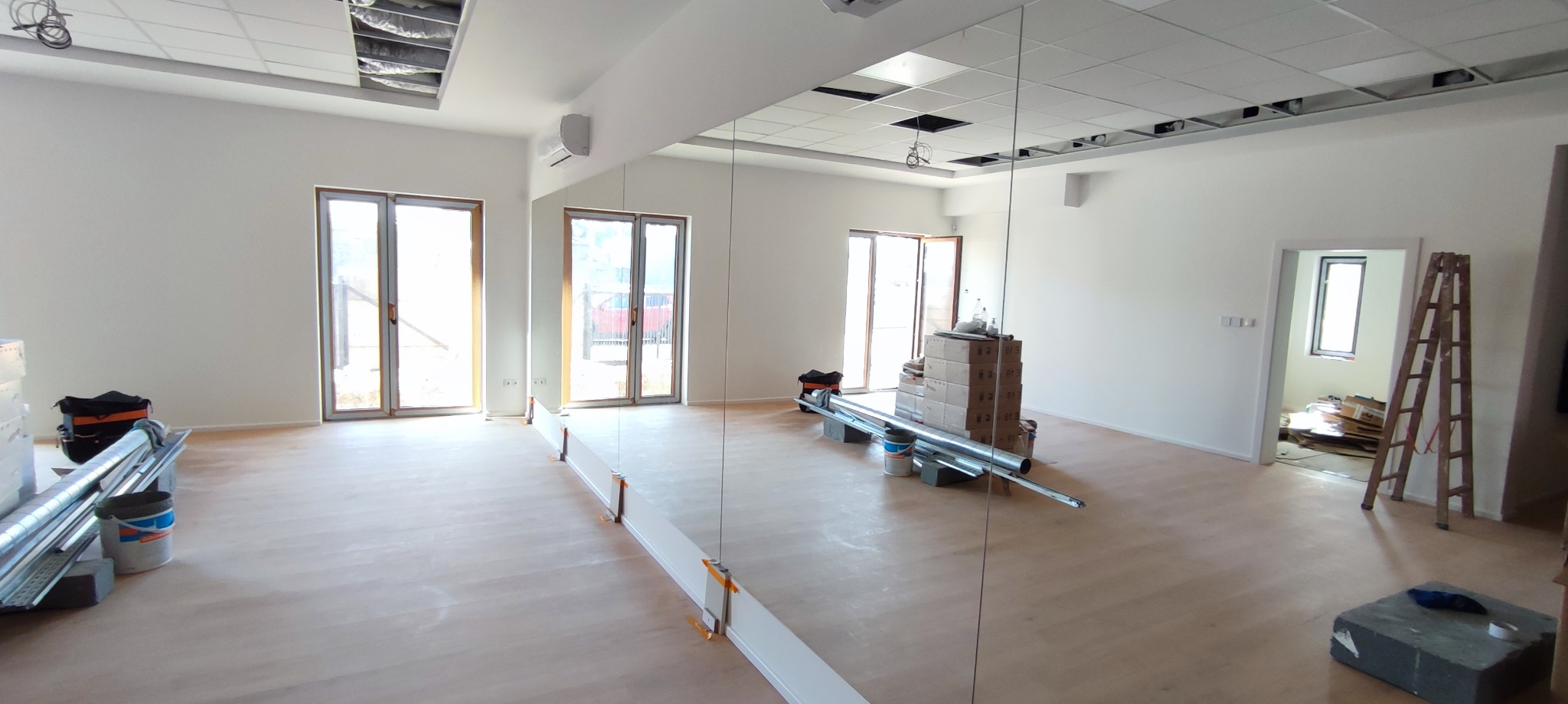 Wnętrze sali z dużym lustrem na ścianie, widoczne materiały budowlane i narzędzia, sugerujące prace wykończeniowe. Jasne ściany i podłoga, okna w tle.