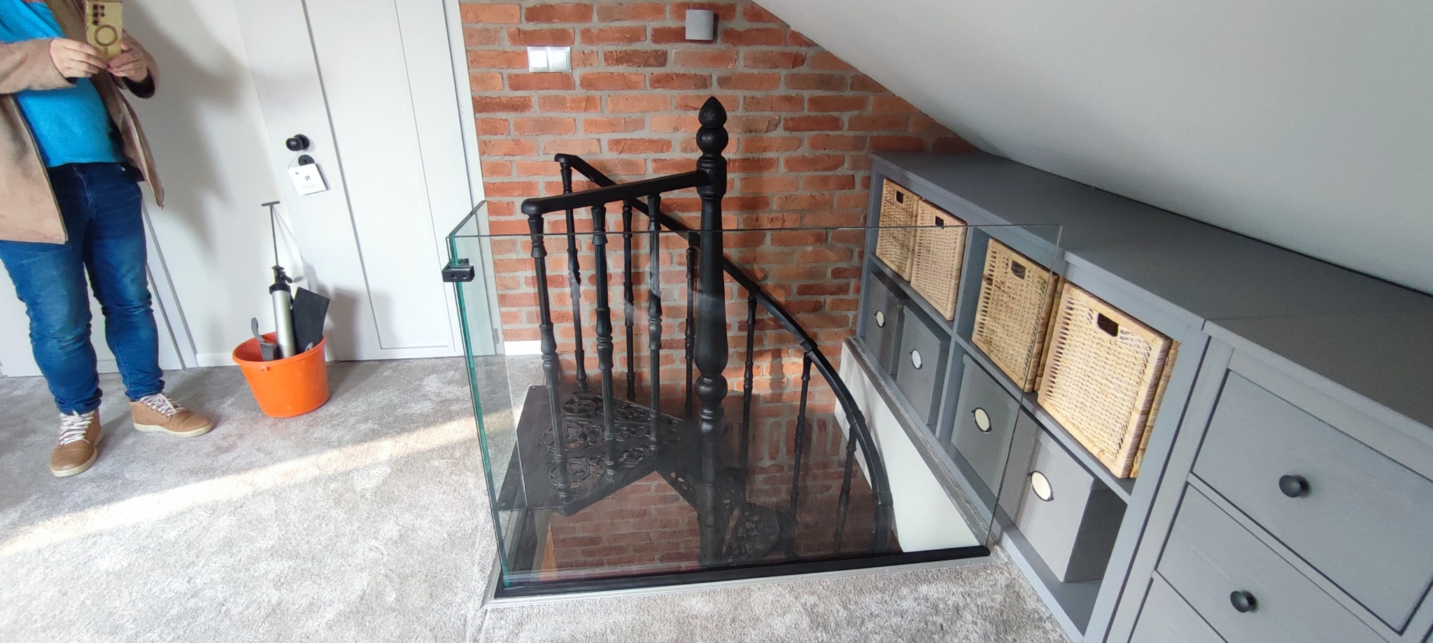 Szklana balustrada zabezpieczająca schody spiralne z czarną, metalową poręczą. W tle ściana z czerwonej cegły i zabudowa meblowa w szarości.