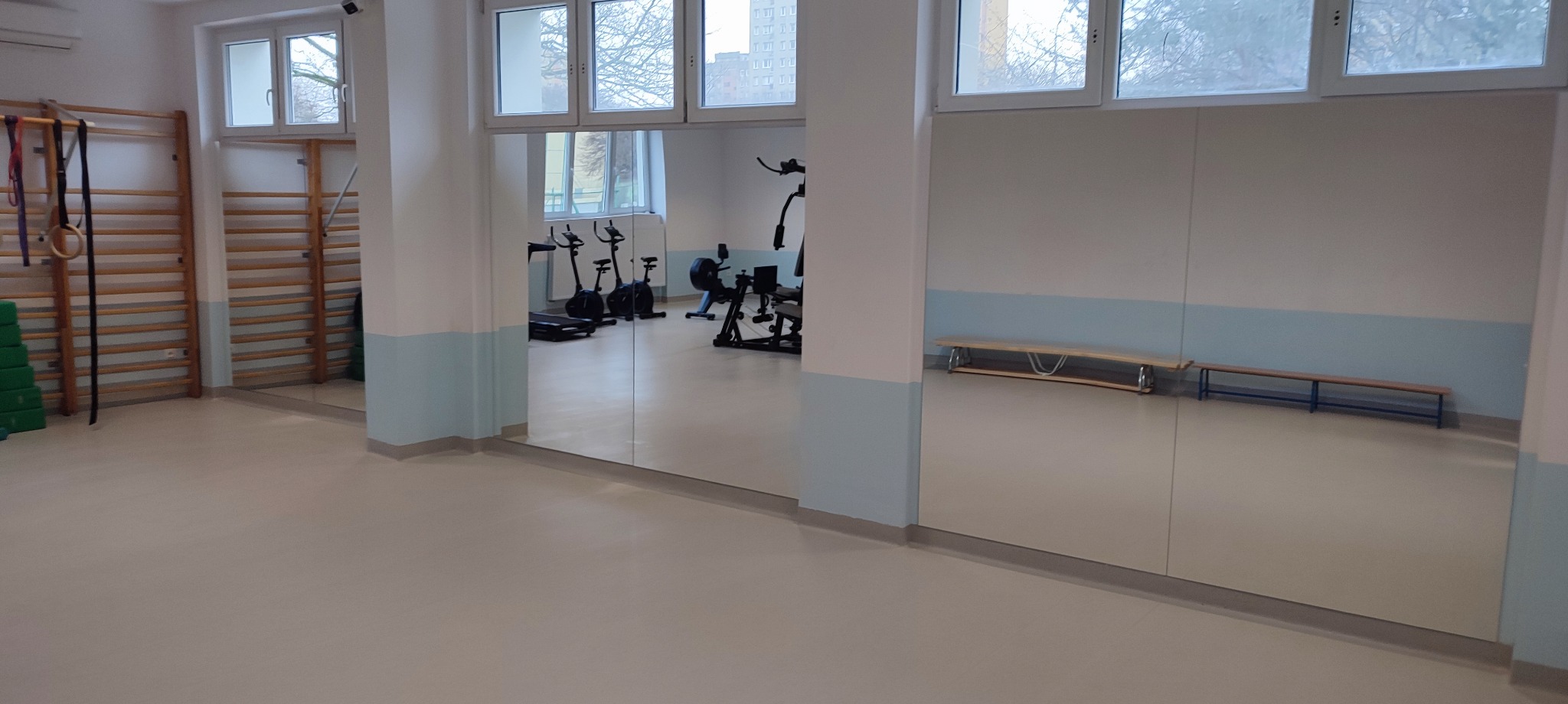 Duże lustra w sali gimnastycznej odbijają sprzęt do ćwiczeń, drabinki gimnastyczne i ławki. Ściany pomalowane w pasy bieli i błękitu. Jasne wnętrze.