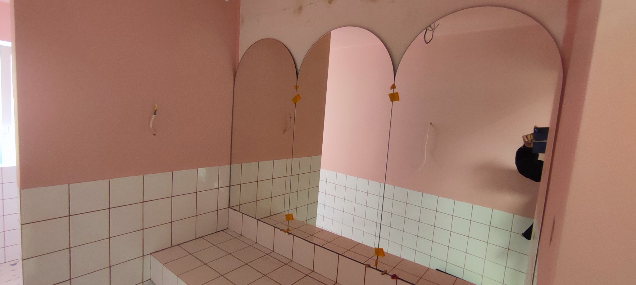 Montaż trzech owalnych luster w łazience z różowymi ścianami i białymi kafelkami. Widoczne przyssawki montażowe i odbicie osoby robiącej zdjęcie.