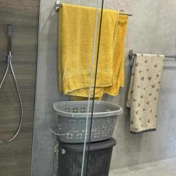 Tomek Usługi Glazurnicze i Wykończenia - Szklana kabina prysznicowa z szarą ścianą, na której wiszą żółte i wzorzyste ręczniki. Pod nimi kosze na pranie, jeden na drugim. Widoczny także panel prysznicowy.