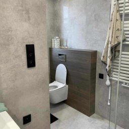 Tomek Usługi Glazurnicze i Wykończenia - Nowoczesna łazienka w Kluczborku z szarymi płytkami, podwieszaną toaletą i grzejnikiem drabinkowym. Ciemne akcenty drewna i minimalistyczne dodatki.