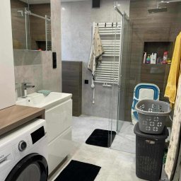 Tomek Usługi Glazurnicze i Wykończenia - Nowoczesna łazienka w szarościach z kabiną prysznicową typu walk-in, pralką i umywalką z szafką. Minimalistyczne czarne dywaniki i akcenty drewniane.