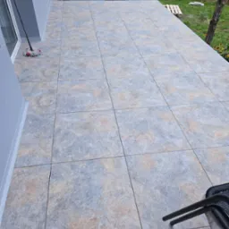 Wyłożony płytkami taras przy domu z mopem w rogu. Szare płytki z beżowymi akcentami. Widok na ogród z paletą i kurami. Część krzeseł w prawym dolnym rogu.