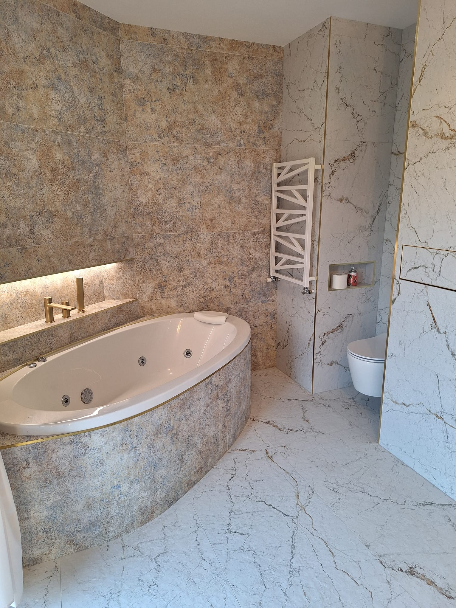 Wykończona łazienka z wanną jacuzzi, grzejnikiem drabinkowym i toaletą, wyłożona płytkami imitującymi marmur w odcieniach beżu i szarości, z subtelnymi złotymi akcentami na krawędziach płytek.