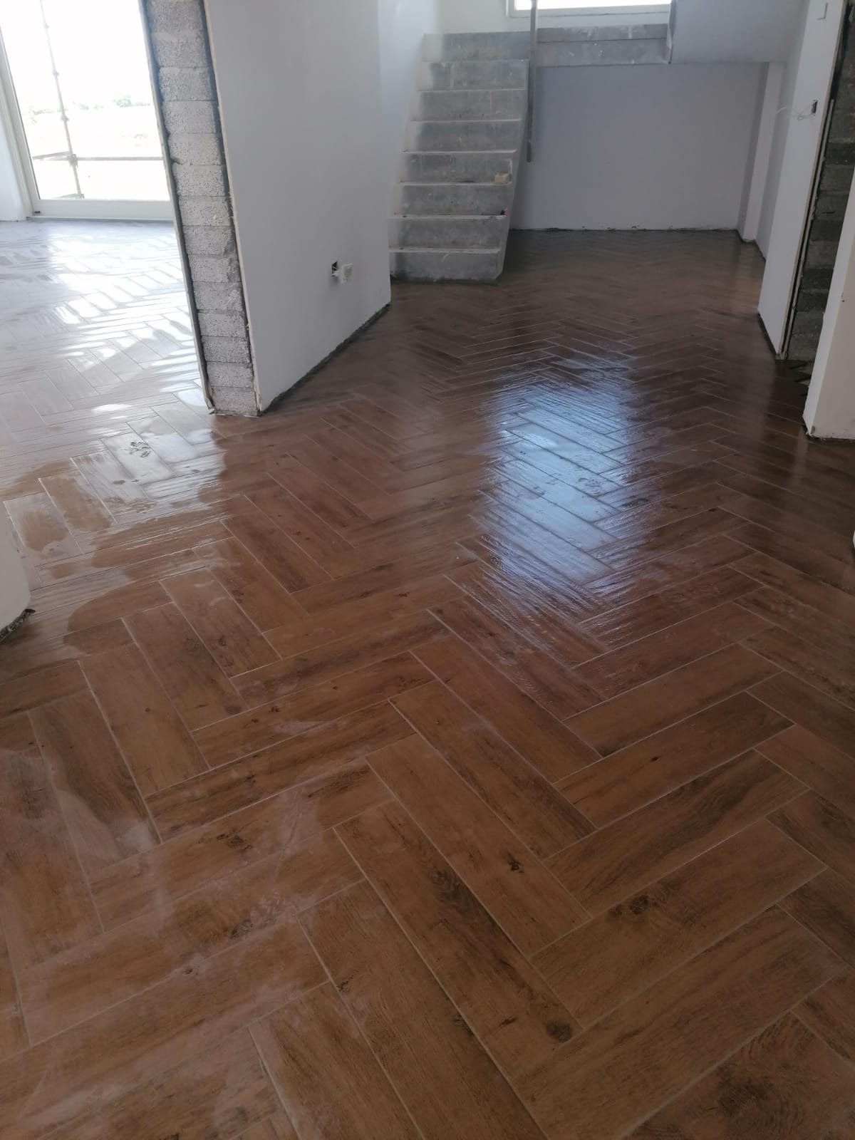 Ułożona w jodełkę, brązowa, błyszcząca podłoga z płytek imitujących drewno w nowym wnętrzu, widoczne schody i fragmenty ścian w stanie surowym.