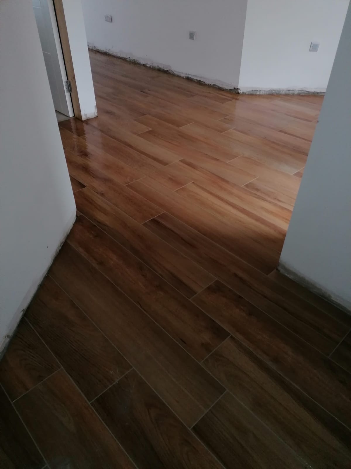 Połyskująca, brązowa podłoga z płytek imitujących drewno ułożona w jodełkę, widoczna w przejściu między pokojami, ściany w stanie surowym wymagają wykończenia.