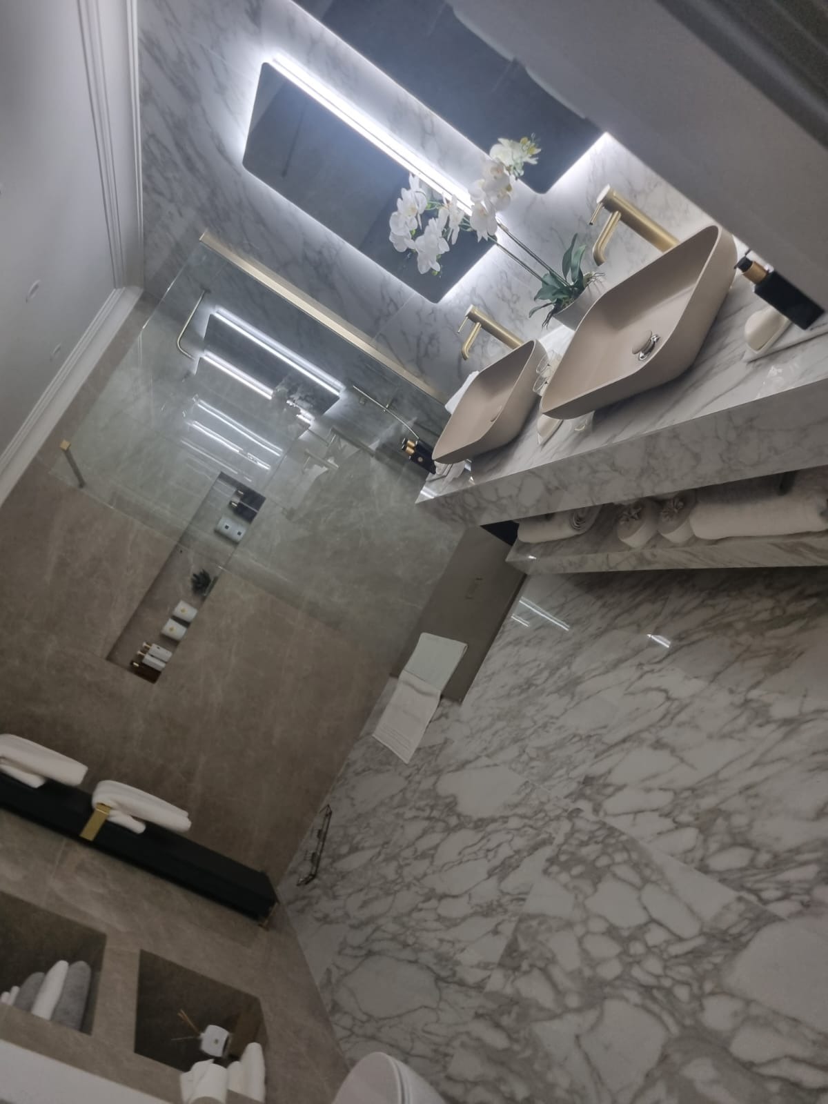 Elegancka łazienka z marmurowymi płytkami, dwiema beżowymi umywalkami nablatowymi z złotymi bateriami, podświetlanym lustrem oraz szklaną kabiną prysznicową z półkami na kosmetyki.