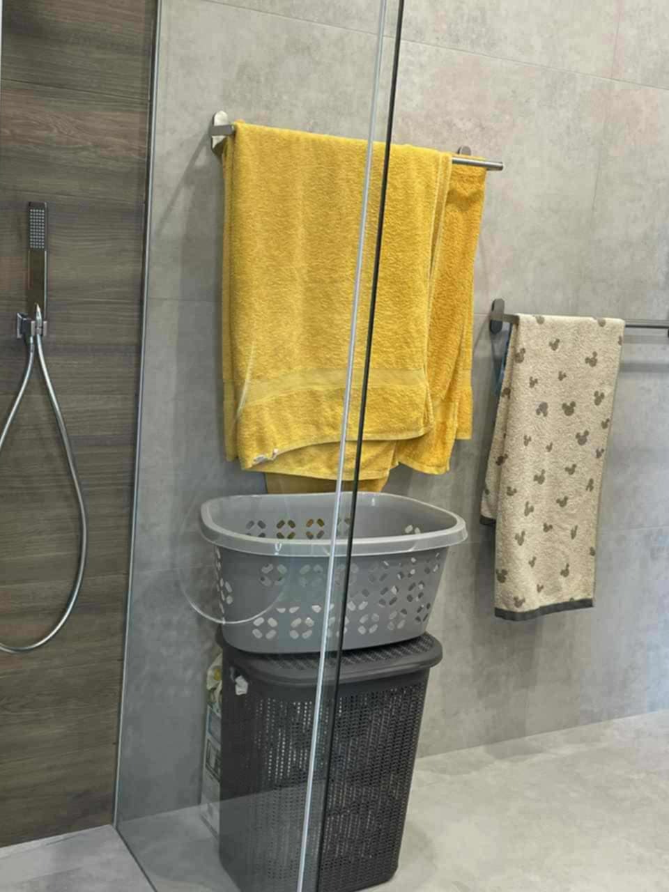 Szklana kabina prysznicowa z szarą ścianą, na której wiszą żółte i wzorzyste ręczniki. Pod nimi kosze na pranie, jeden na drugim. Widoczny także panel prysznicowy.