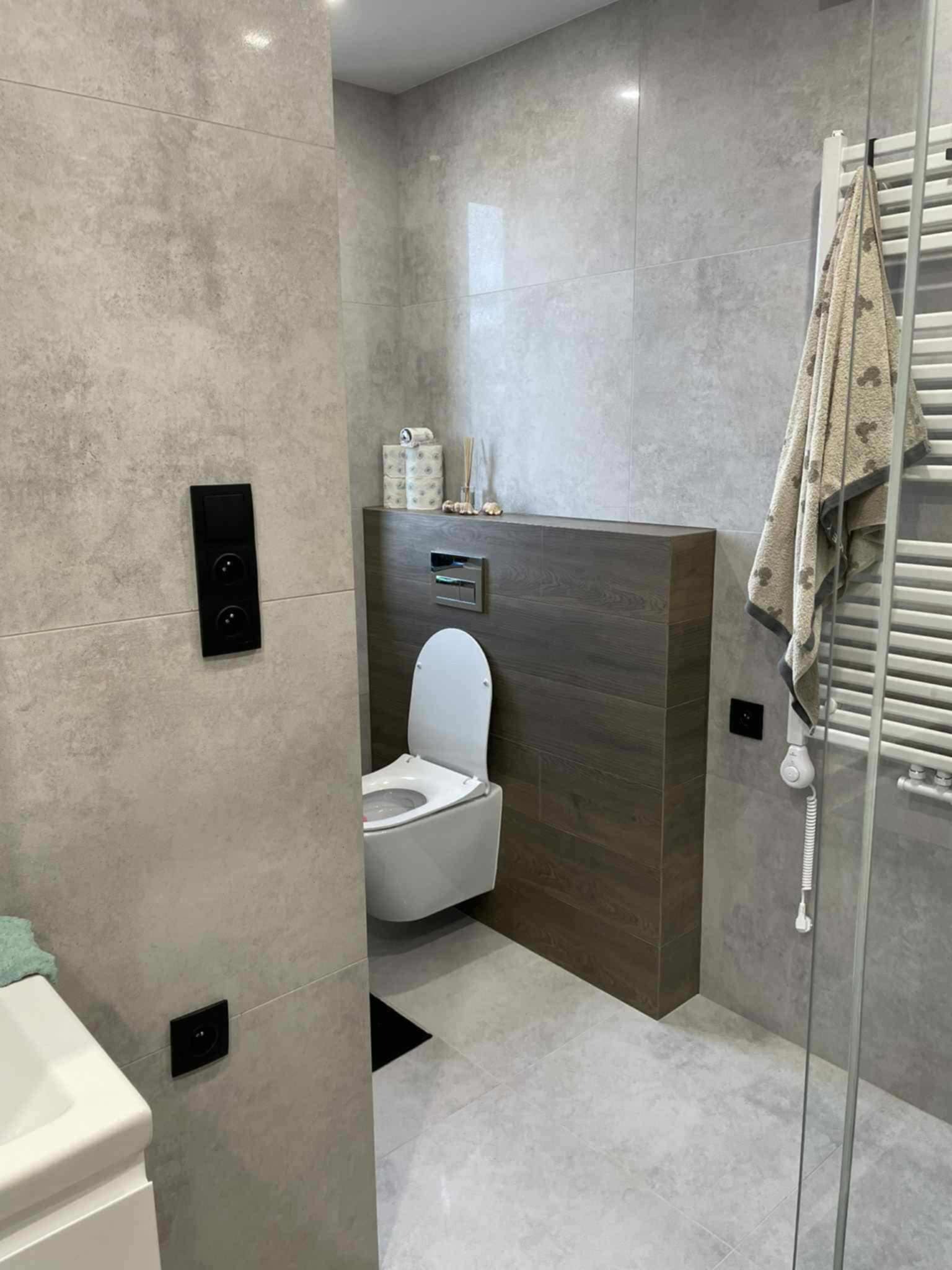 Nowoczesna łazienka w Kluczborku z szarymi płytkami, podwieszaną toaletą i grzejnikiem drabinkowym. Ciemne akcenty drewna i minimalistyczne dodatki.