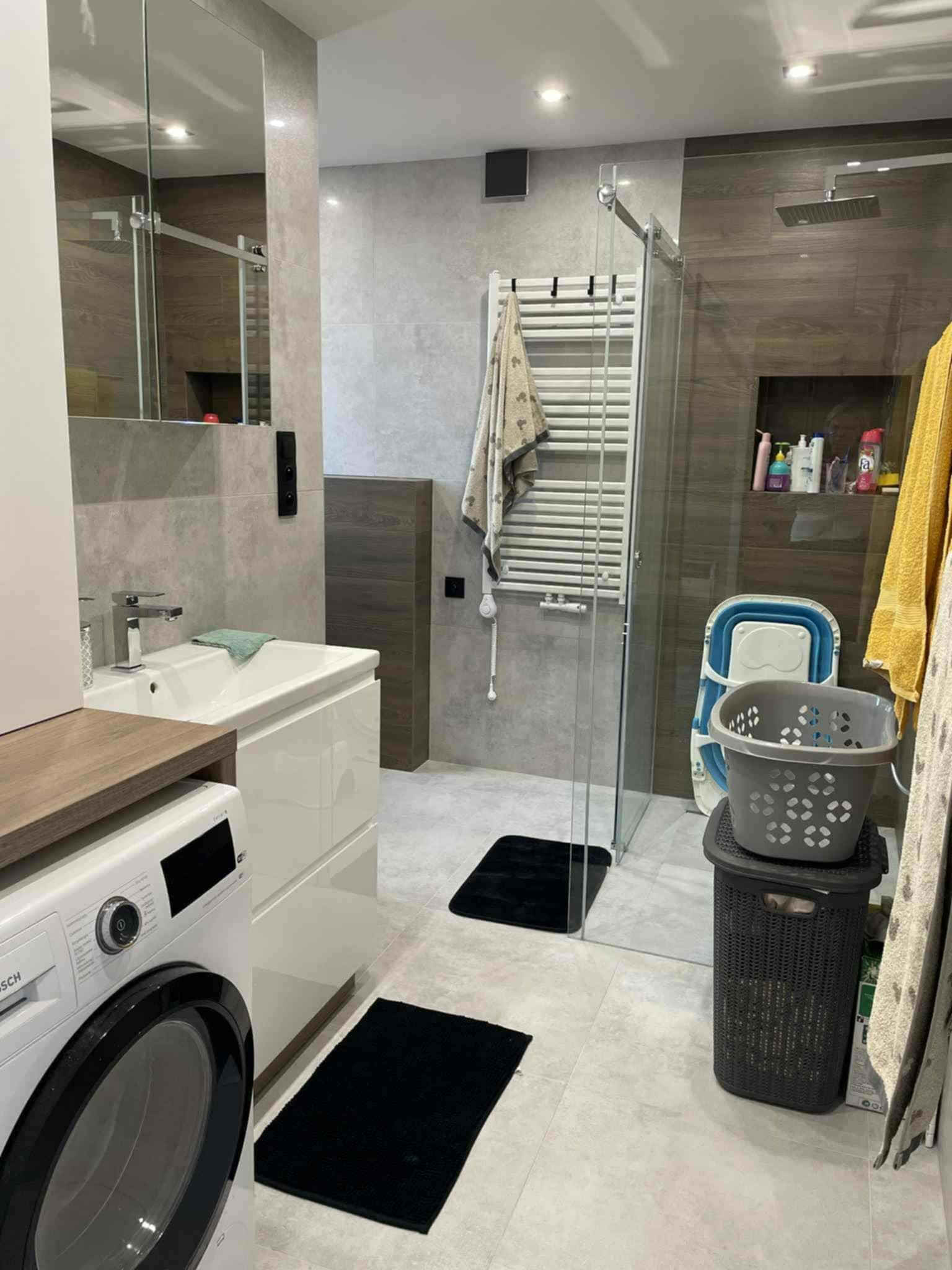 Nowoczesna łazienka w szarościach z kabiną prysznicową typu walk-in, pralką i umywalką z szafką. Minimalistyczne czarne dywaniki i akcenty drewniane.