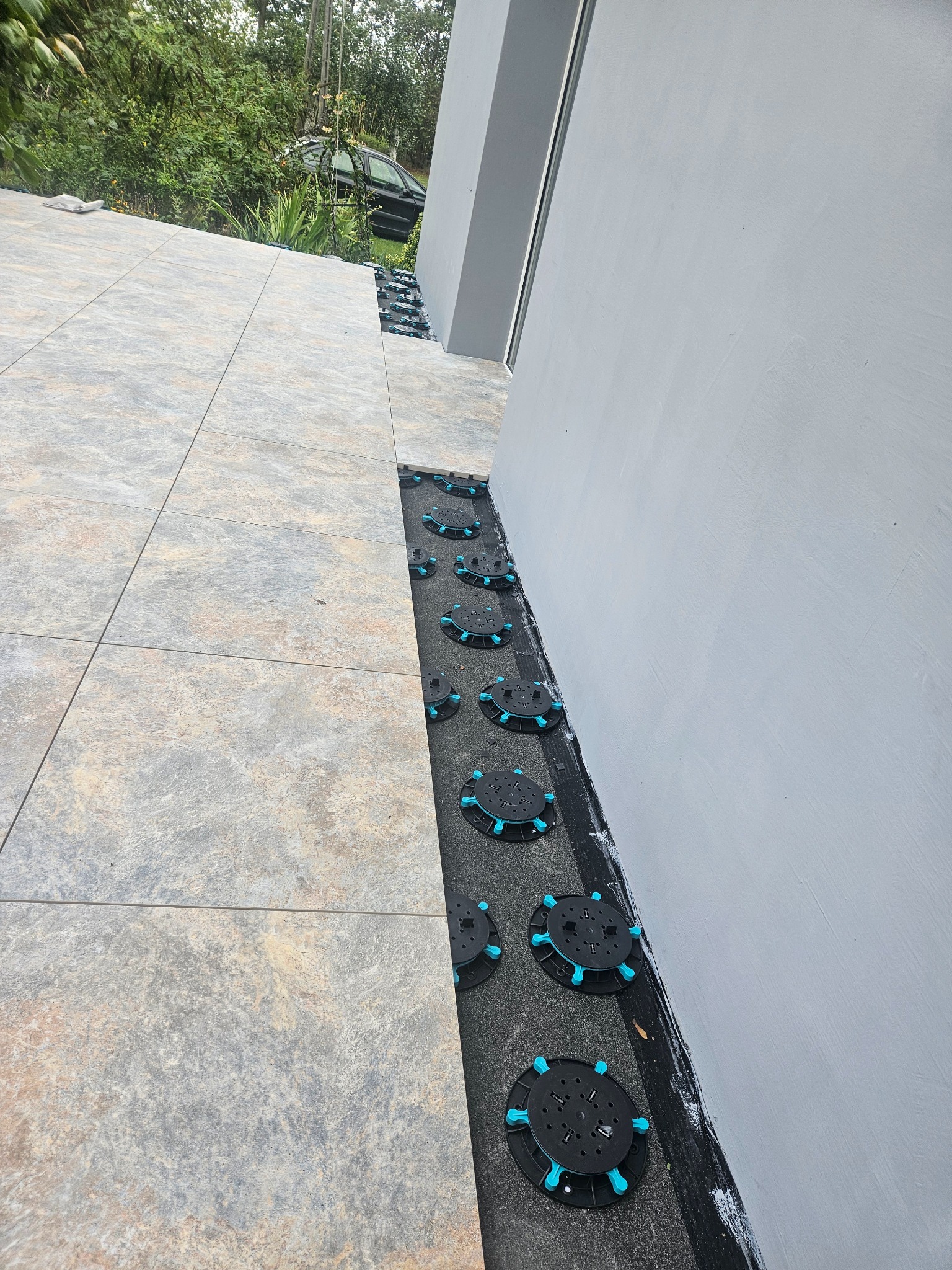 Częściowo ułożone płytki tarasowe na regulowanych wspornikach z tworzywa sztucznego, widoczne tło z zielenią i fragmentem samochodu.