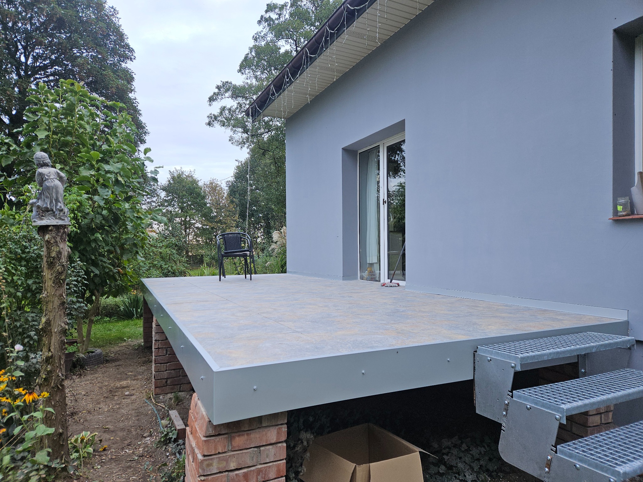 Wykończony taras z szarymi płytkami i metalowymi schodami. Widoczne ceglane podpory, krzesło na tarasie i fragment budynku z oknem. Oświetlenie zewnętrzne.