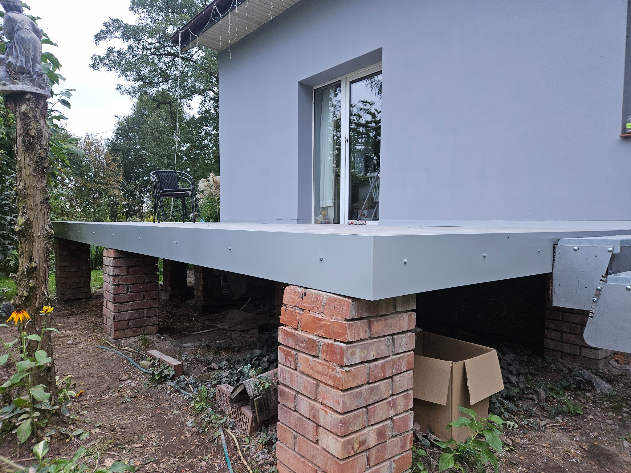 Szary taras na ceglanych podporach przy domu, widok z boku. Na tarasie krzesło i akcesoria. Częściowo widoczne drzewa i roślinność w tle.