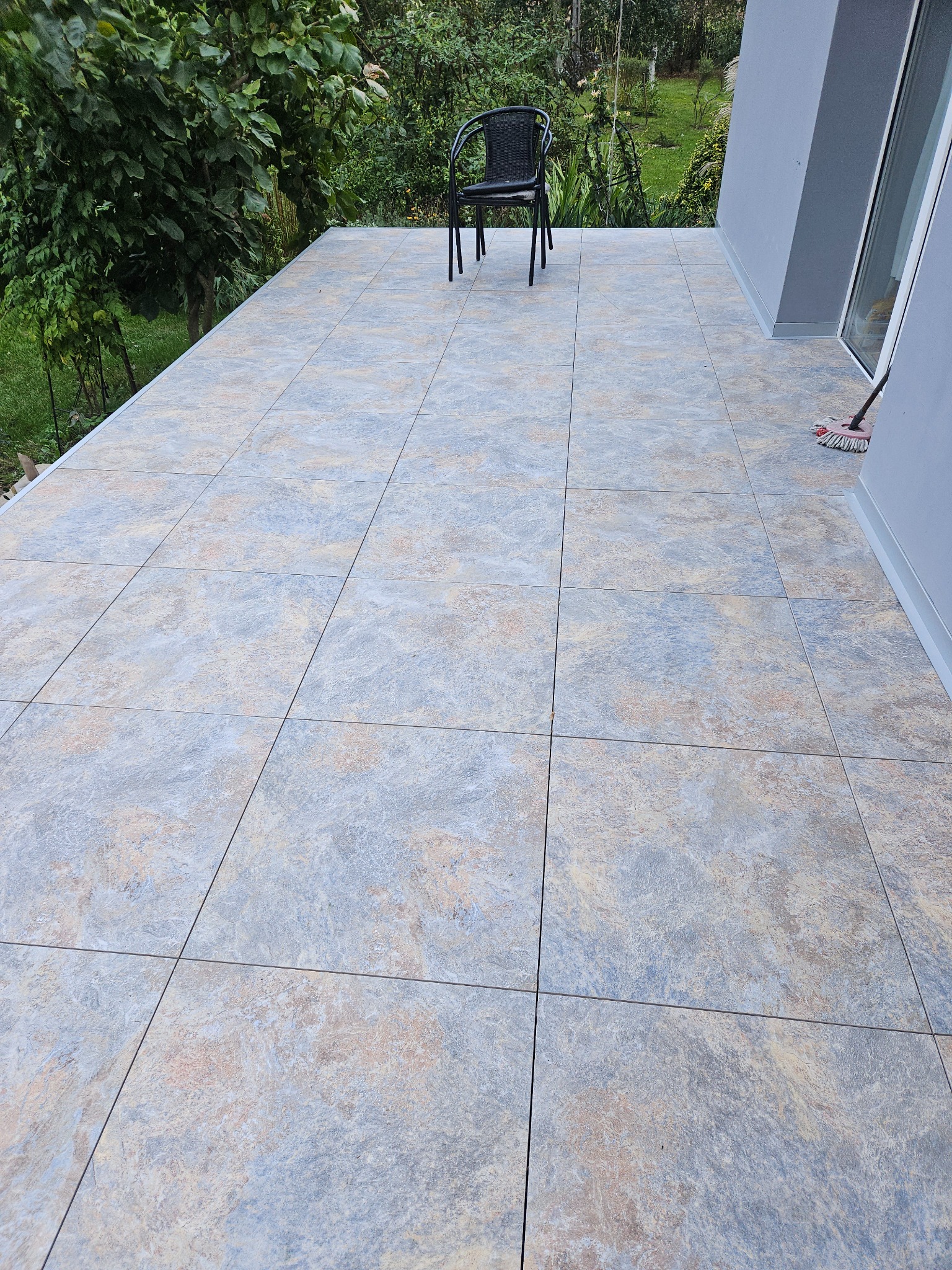 Wykończony taras z szaro-beżowych płytek, na którym stoi czarne krzesło. W tle zieleń ogrodu. Widoczny fragment ściany budynku z oknem.