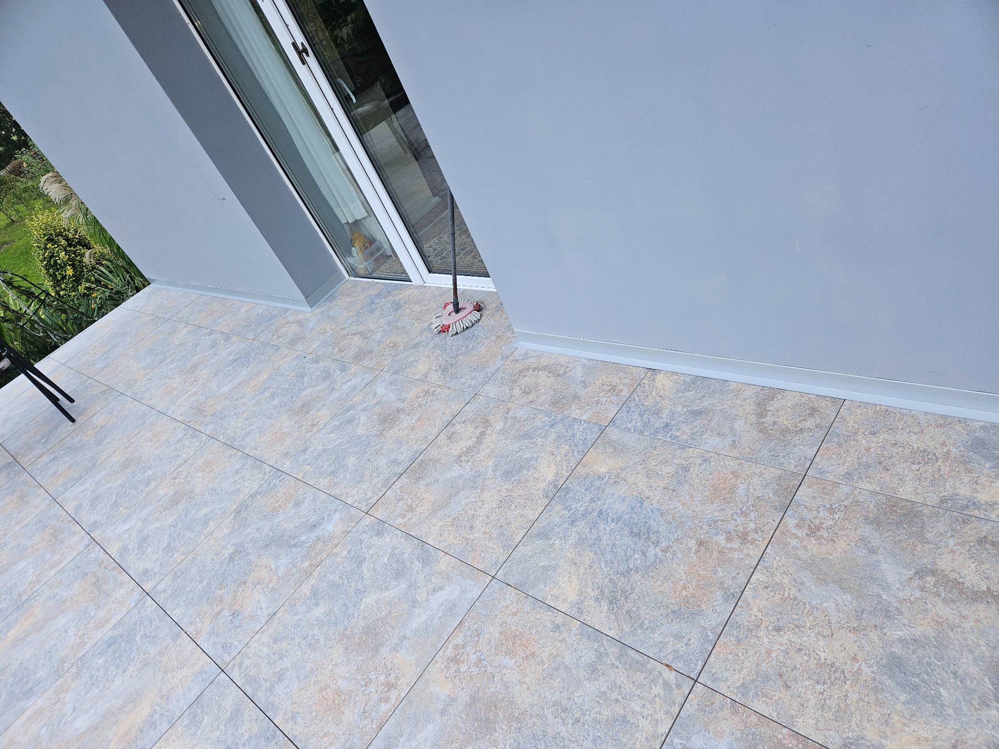 Kafelkowany balkon z szarymi ścianami i mopem w rogu. Płytki w odcieniach beżu i szarości. Widok z góry na wykończony taras z widocznymi fugami i detalami wykończeniowymi.